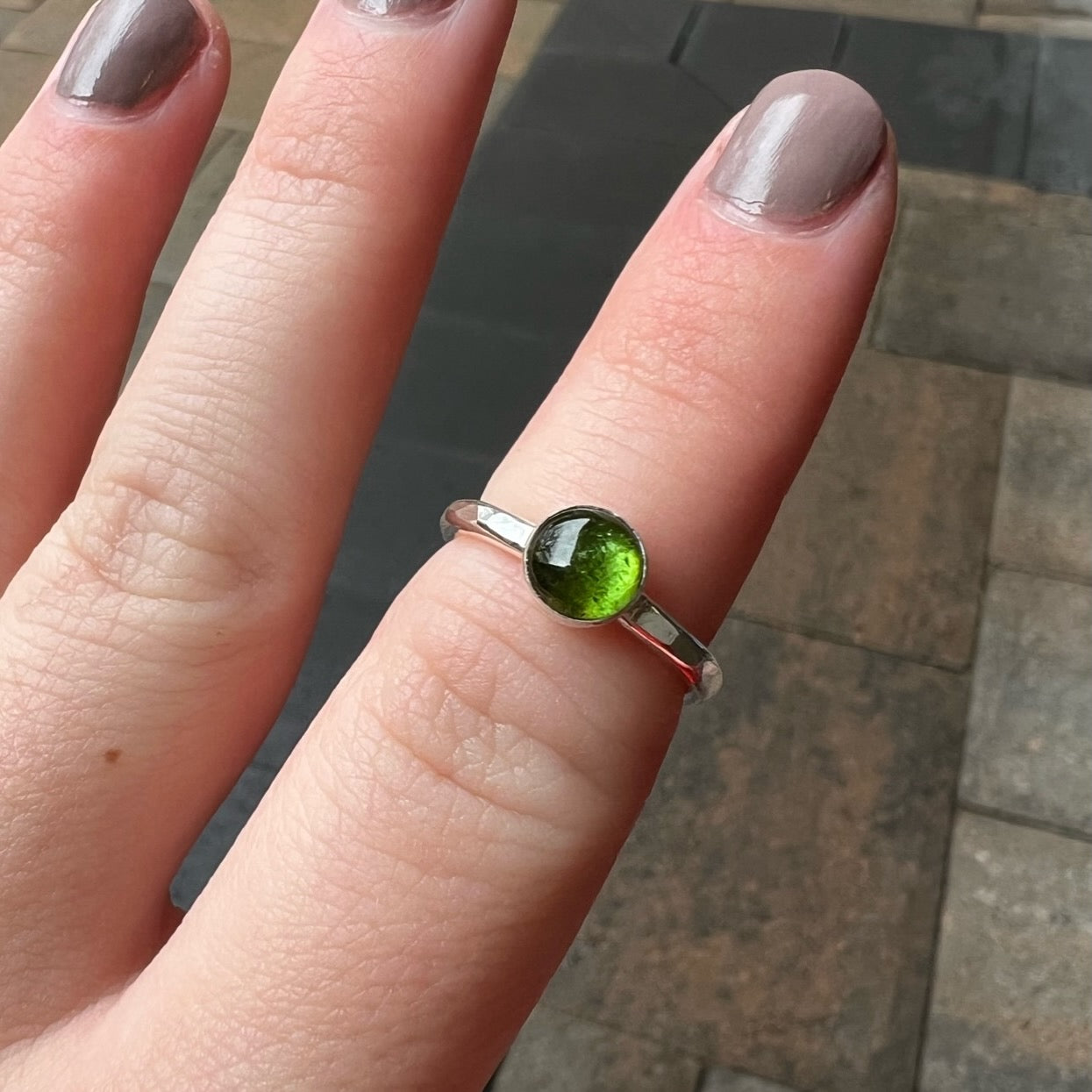 A Round 6mm green tourmaline gemstone set in a sterling silver bezel setting on a sturdy hammered band modeled on a finger. 