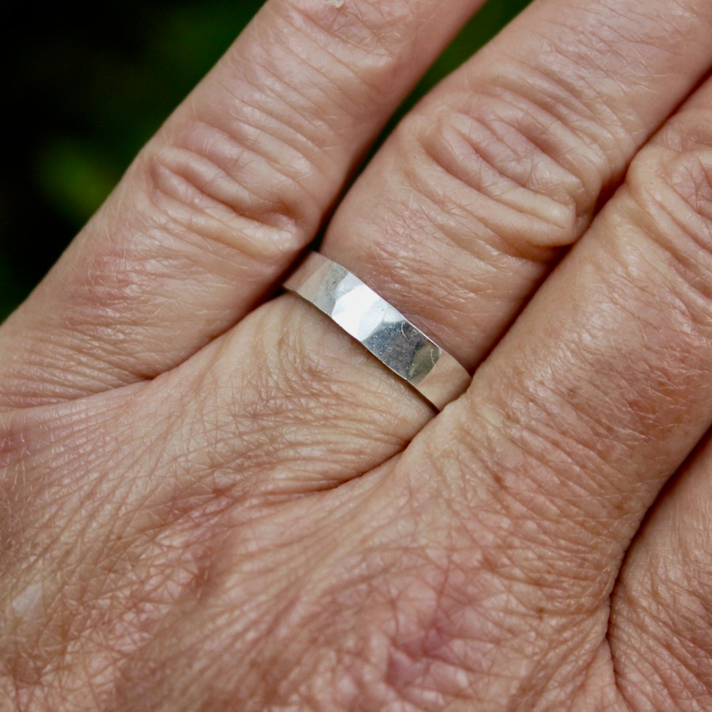 A handmade 4mm wide x 1mm thick flat sterling silver band with a hammered texture and bright finish shown on a ring finger. 