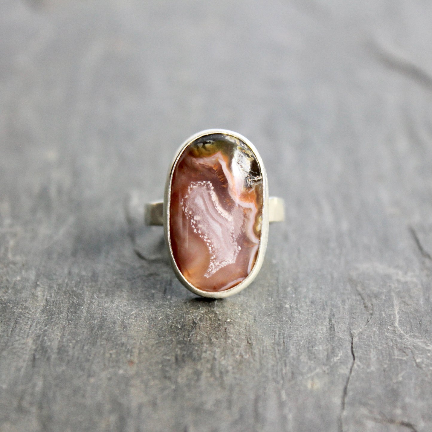 Silver ring with an oval stone on a gray surface.T his pretty ring has an oval oblong pinkish Mexican agua nueva agate cabochon set in a fine and sterling silver bezel setting on a sturdy hammered band. Size 6 Handmade by Will Macy in Corvallis, OR.