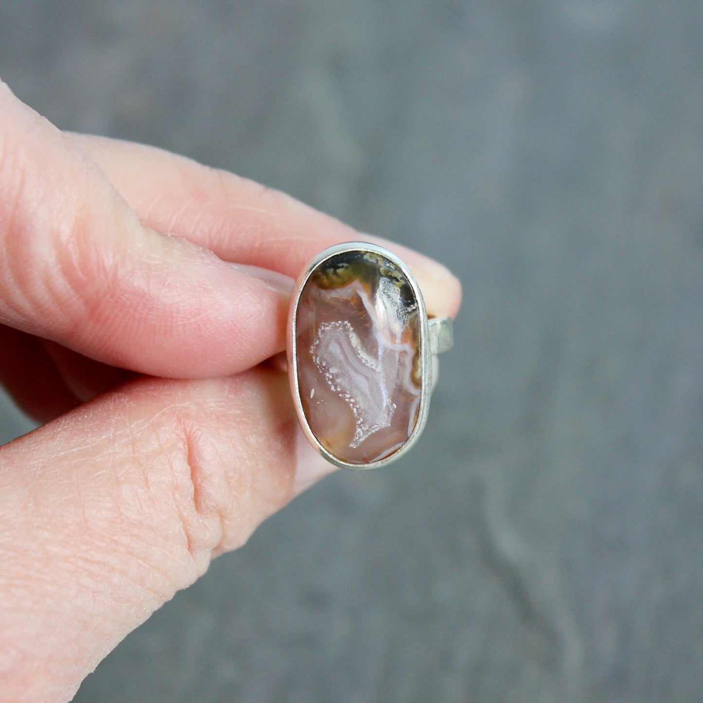 Hand holding a ring with an agate stone against a gray background.This pretty ring has an oval oblong pinkish Mexican aqua nueva agate cabochon set in a fine and sterling silver bezel setting on a sturdy hammered band. Size 6 Handmade by Will Macy in Corvallis, OR.