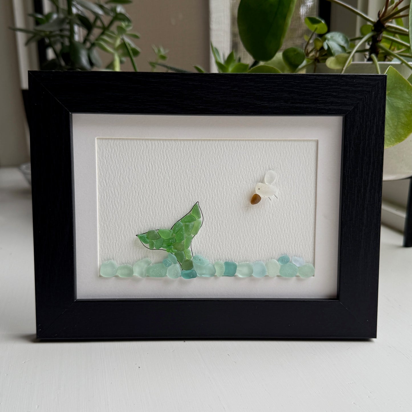Framed artwork with a green mermaid tail and small white flower on a textured white background.
