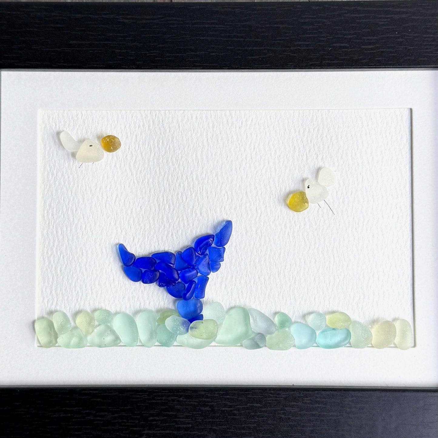 Artistic depiction of a whale tail made from small stones on a white background with a black frame.