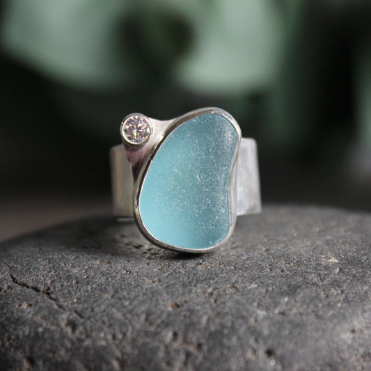This statement ring has a large piece of aqua blue sea glass set in a fine and sterling silver bezel setting with a 4mm tube set cubic zirconia on a 1/2 inch wide sterling silver band. 