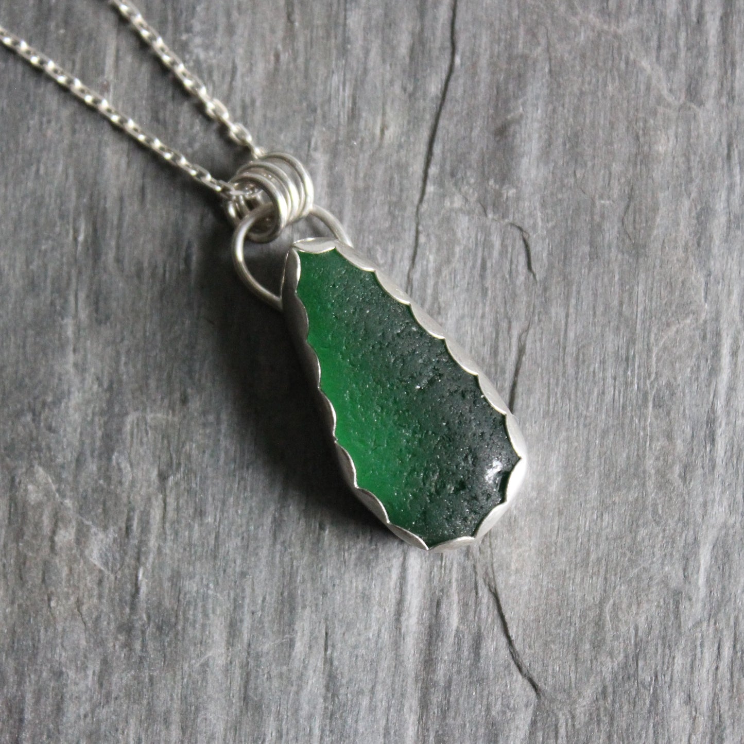 A large dark emerald green piece of sea glass set in a fine and sterling silver scalloped bezel setting with a sterling silver chain. 