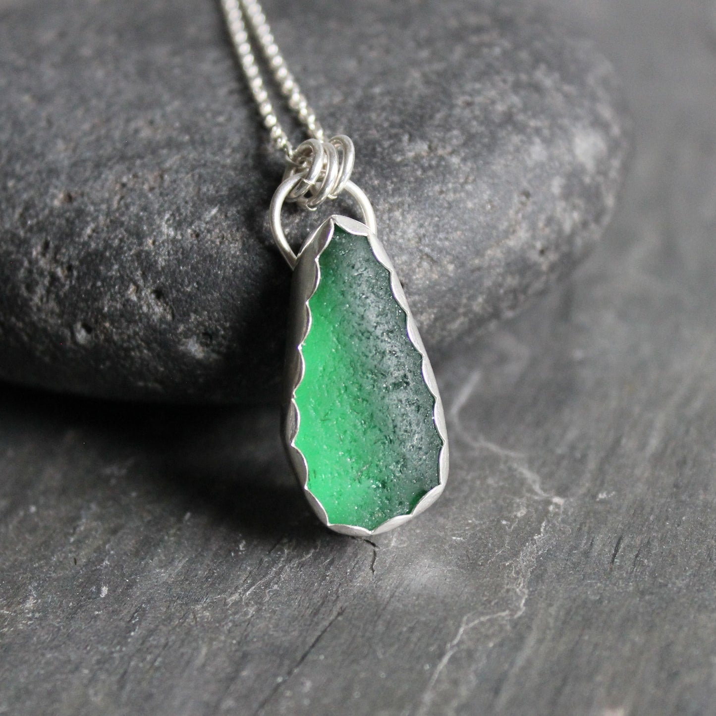 A large dark emerald green piece of sea glass set in a fine and sterling silver scalloped bezel setting with a sterling silver chain. 