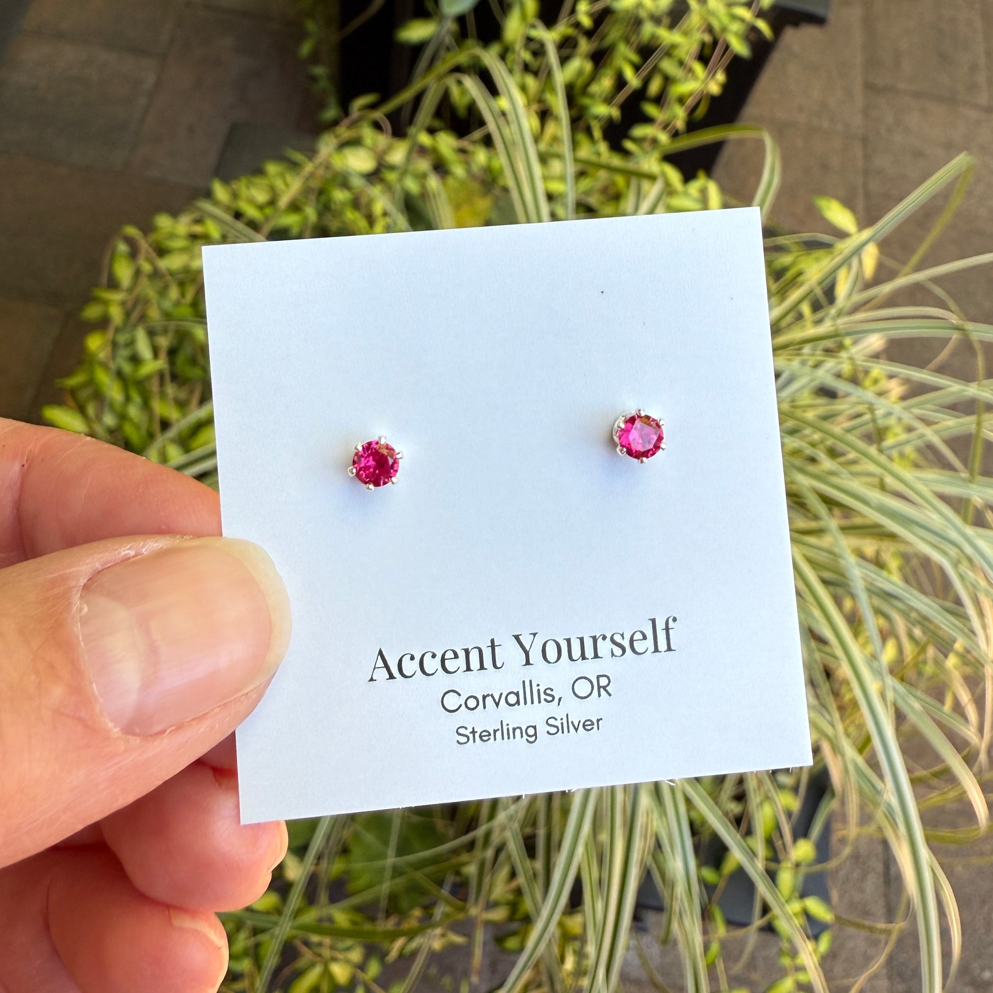 Pink stud earrings on a card held by a hand with plants in the background