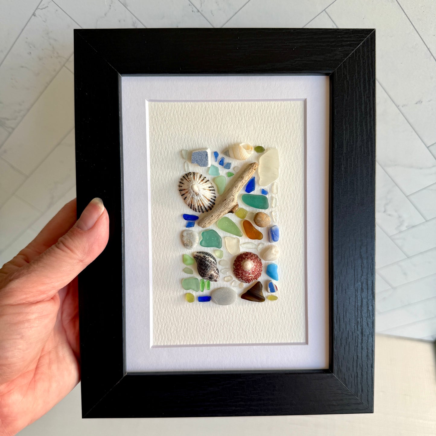 Framed artwork with sea glass and shells held by a hand on a light background
