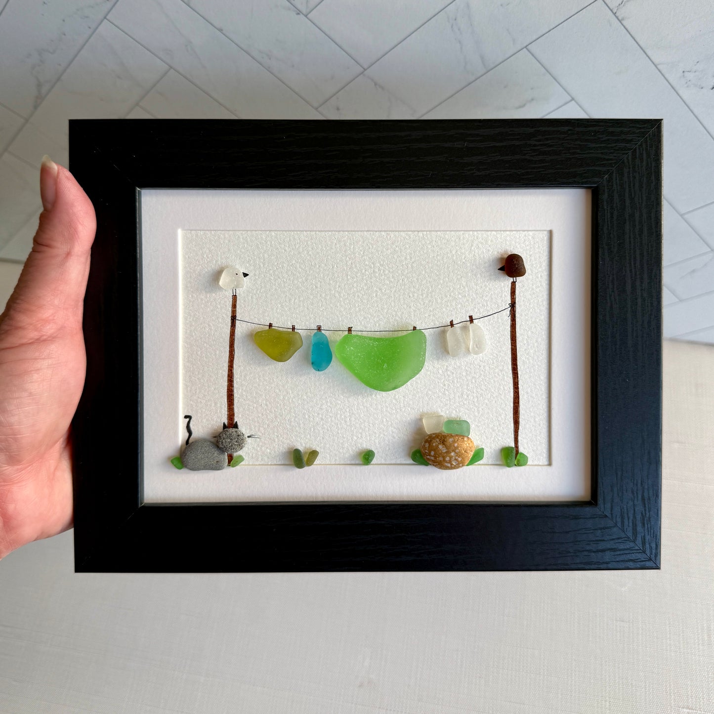 Framed artwork of a cat watching clothes dry on a line with various colored stones.