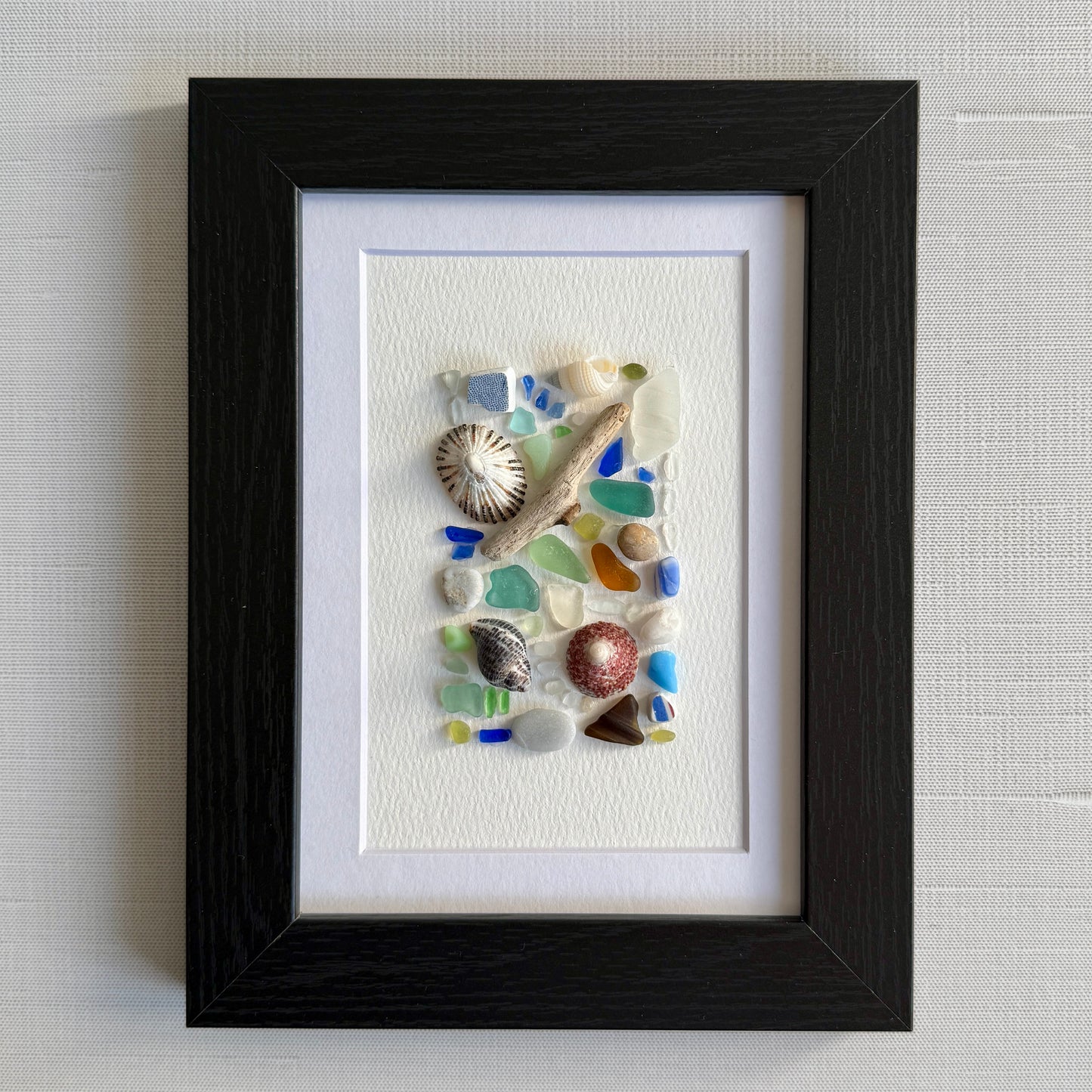Framed artwork with seashells and sea glass pieces on a textured wall background