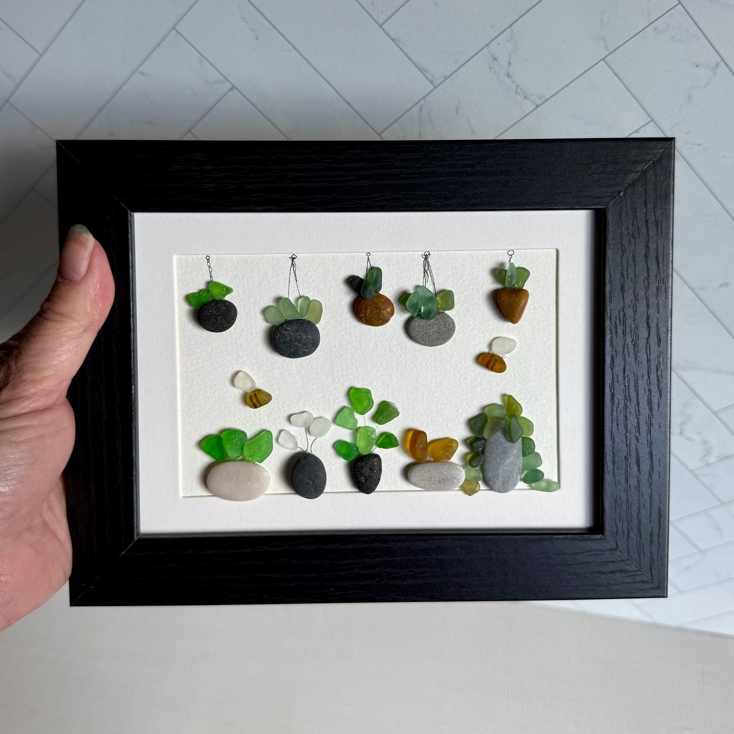 Framed artwork of small plants made from stones and sea glass on a white background