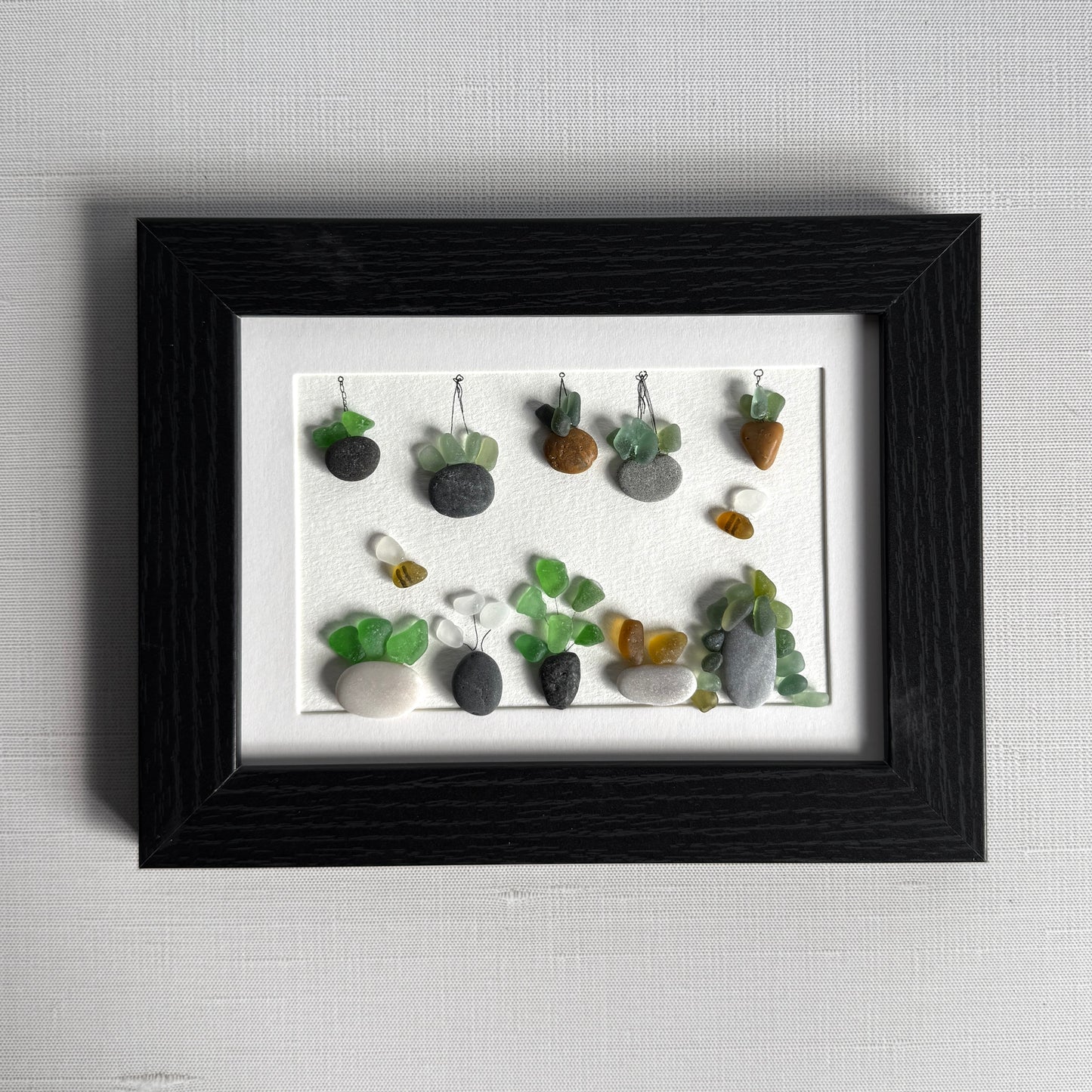 Framed artwork of small plants made from stones and sea glass on a white background