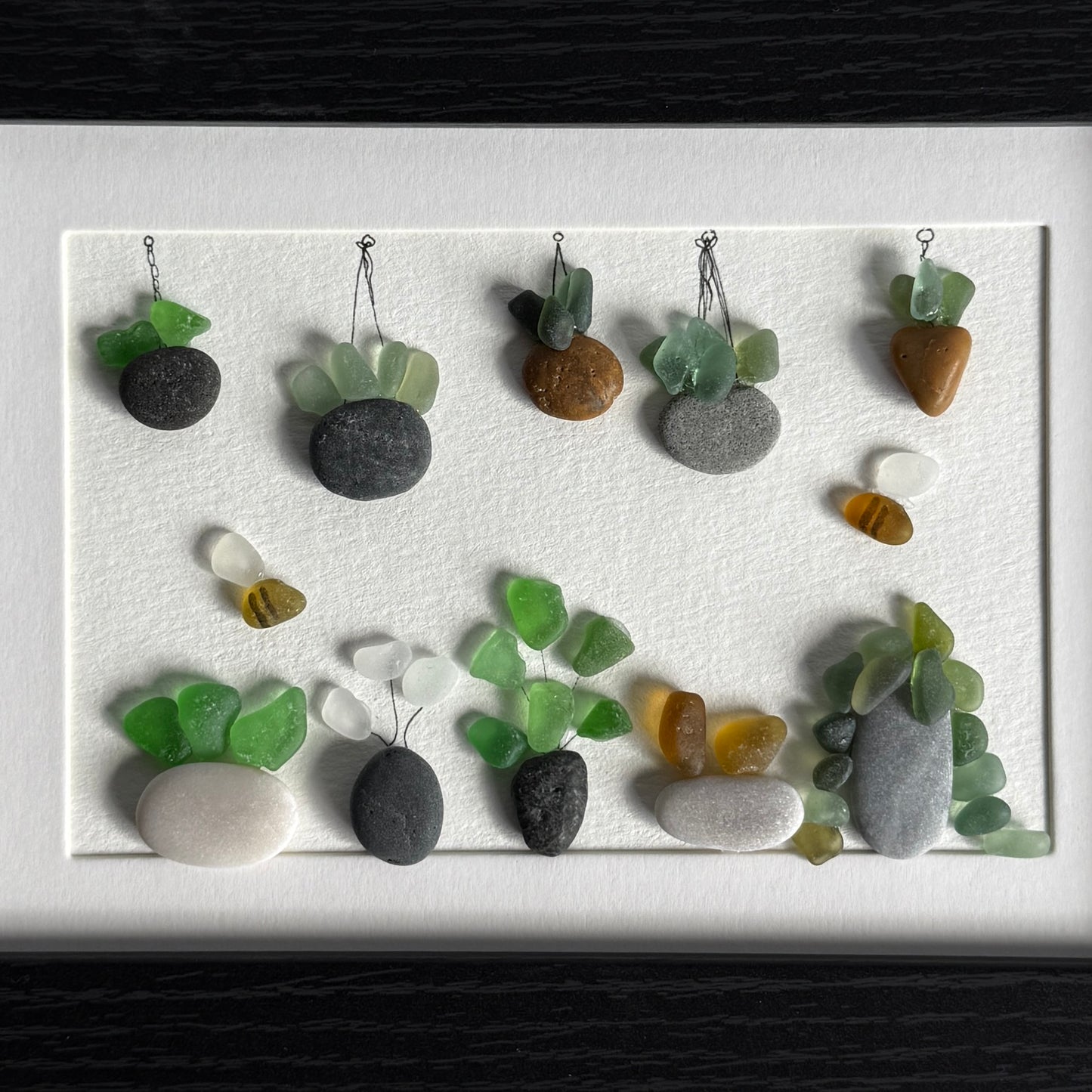 Framed artwork of small plants made from stones and sea glass on a white background