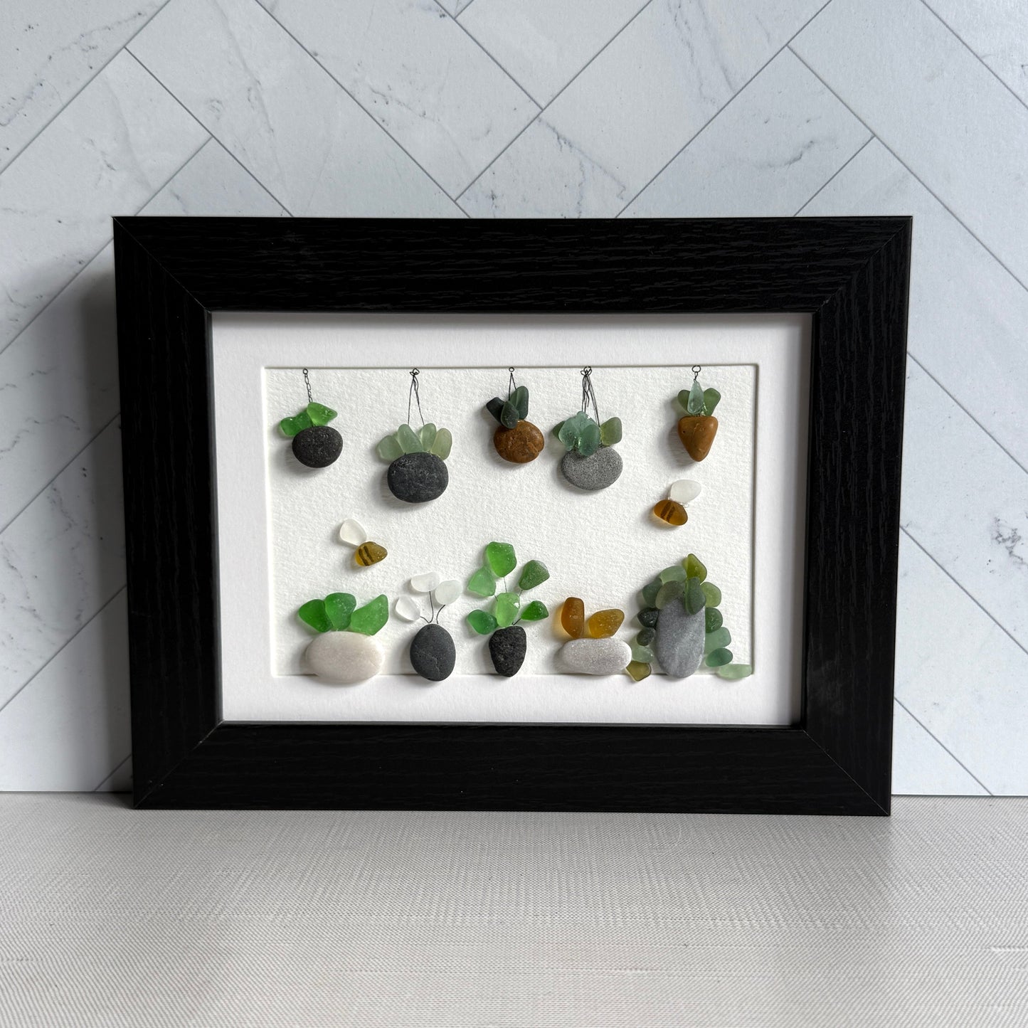 Framed artwork of small plants made from stones and sea glass on a white background