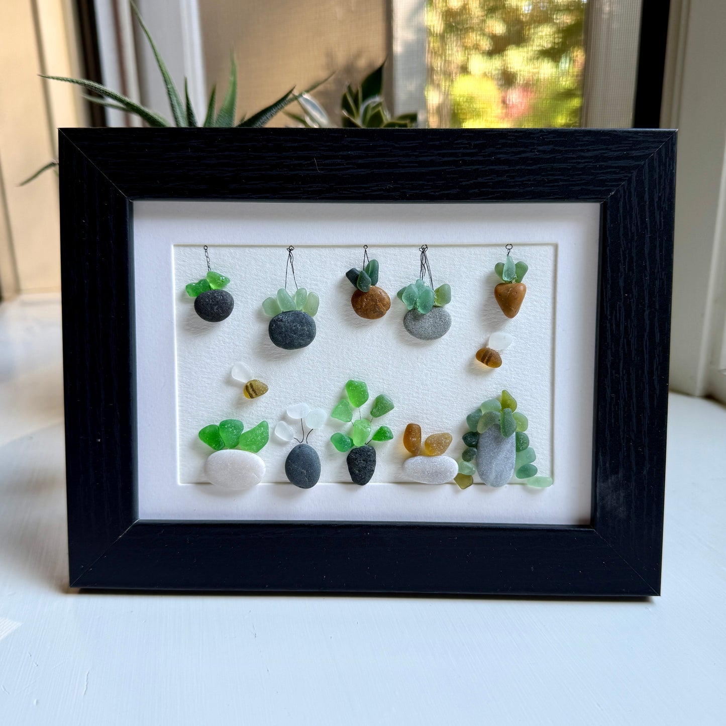 Framed artwork of small plants made from stones and sea glass on a white background