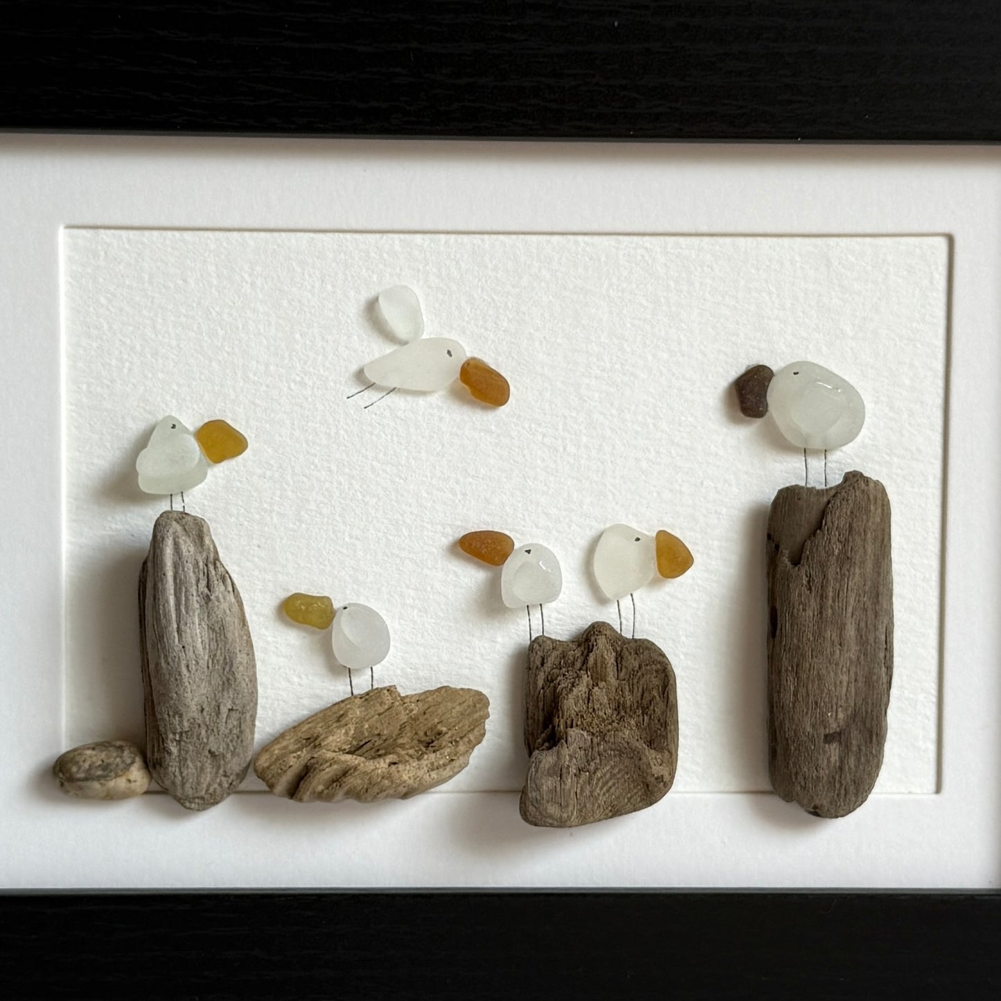 Framed sea glass artwork made of sea glass and driftwood on a white surface.