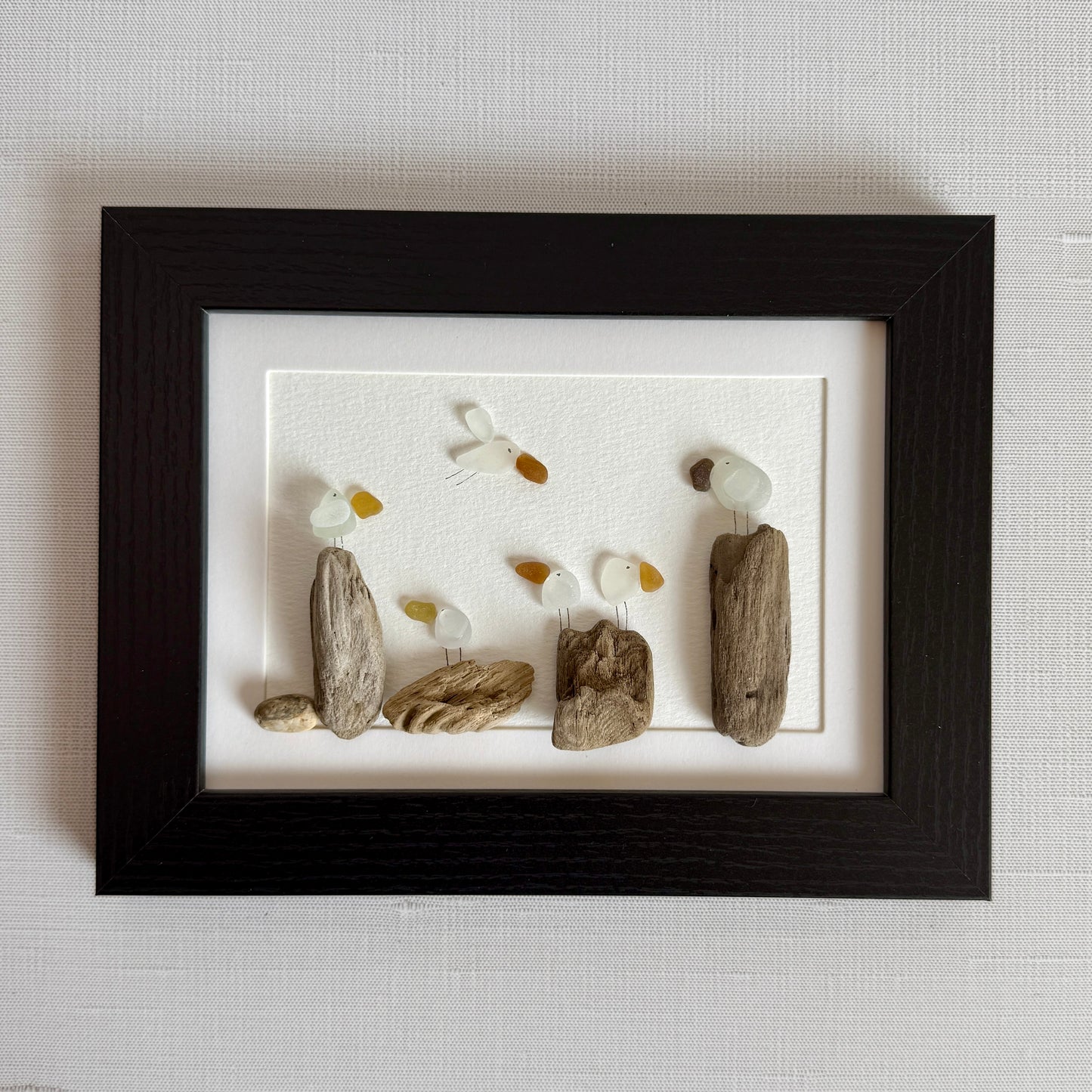 Framed sea glass artwork made of sea glass and driftwood on a white surface.