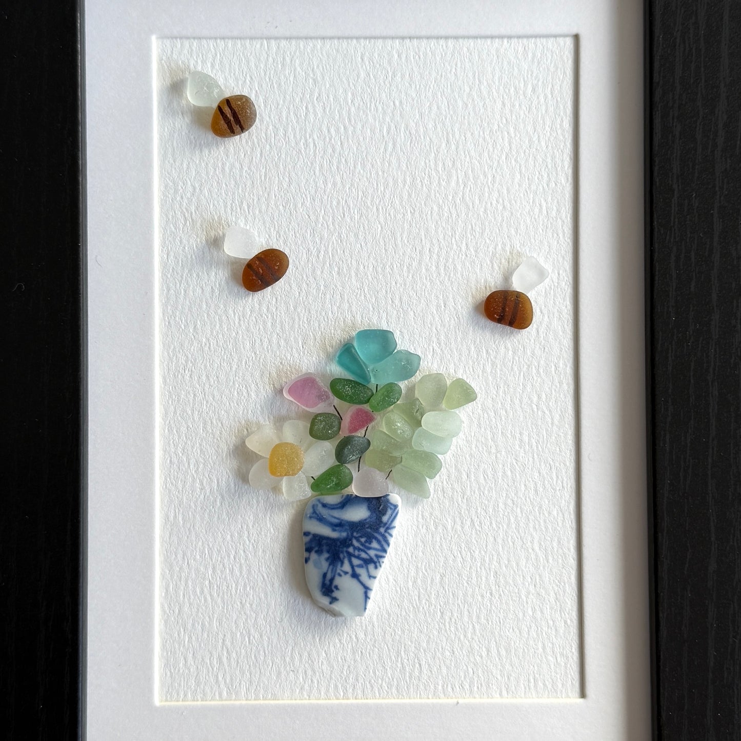 Framed artwork made from colorful sea glass shaped like a flower with a blue and white vase.