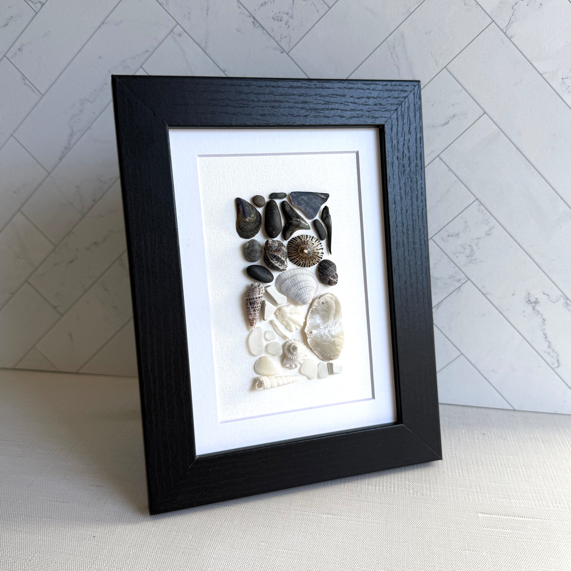 Framed artwork of pebbles, shells and sea glass on a white background