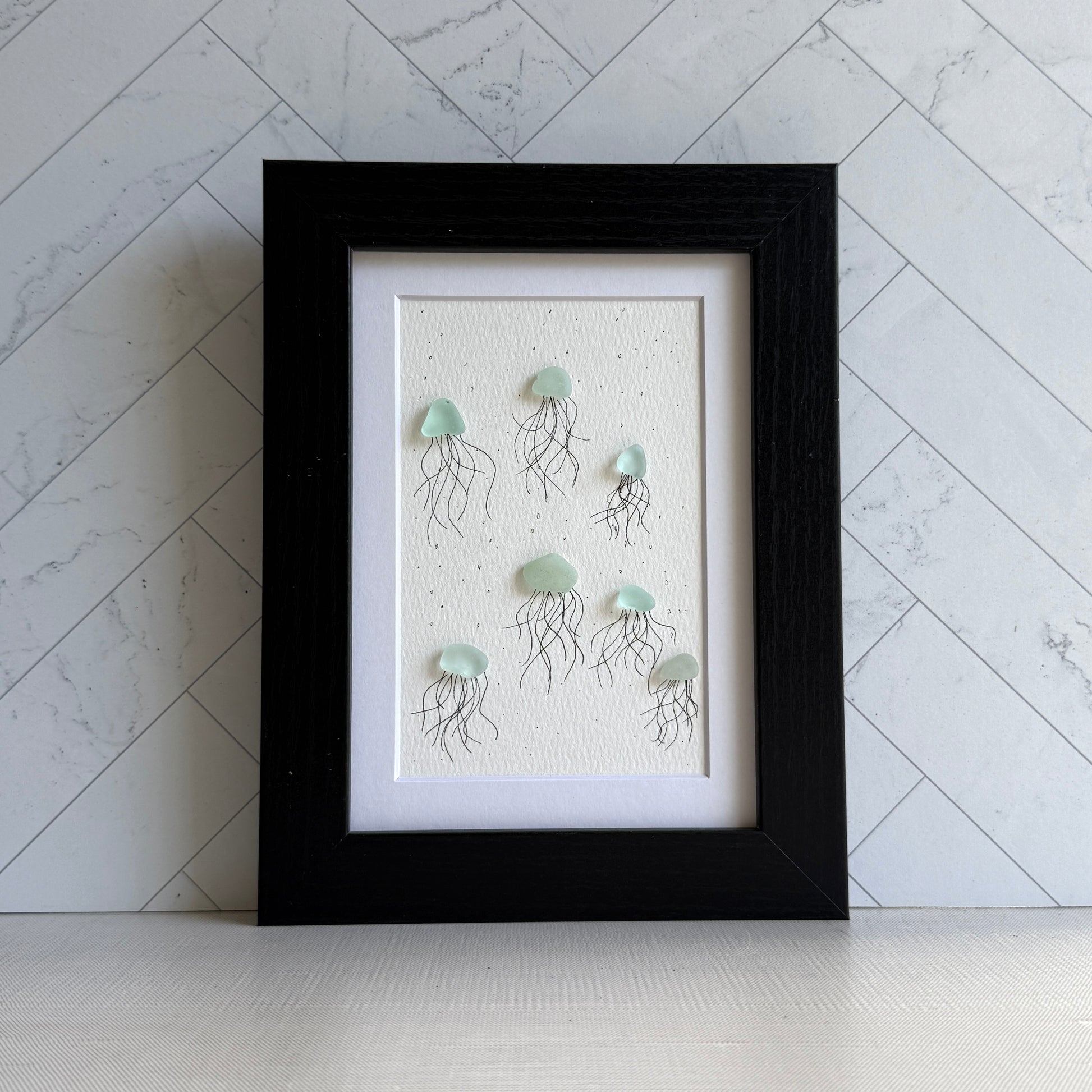 Framed sea glass artwork of jellyfish on a textured white background with a black frame.