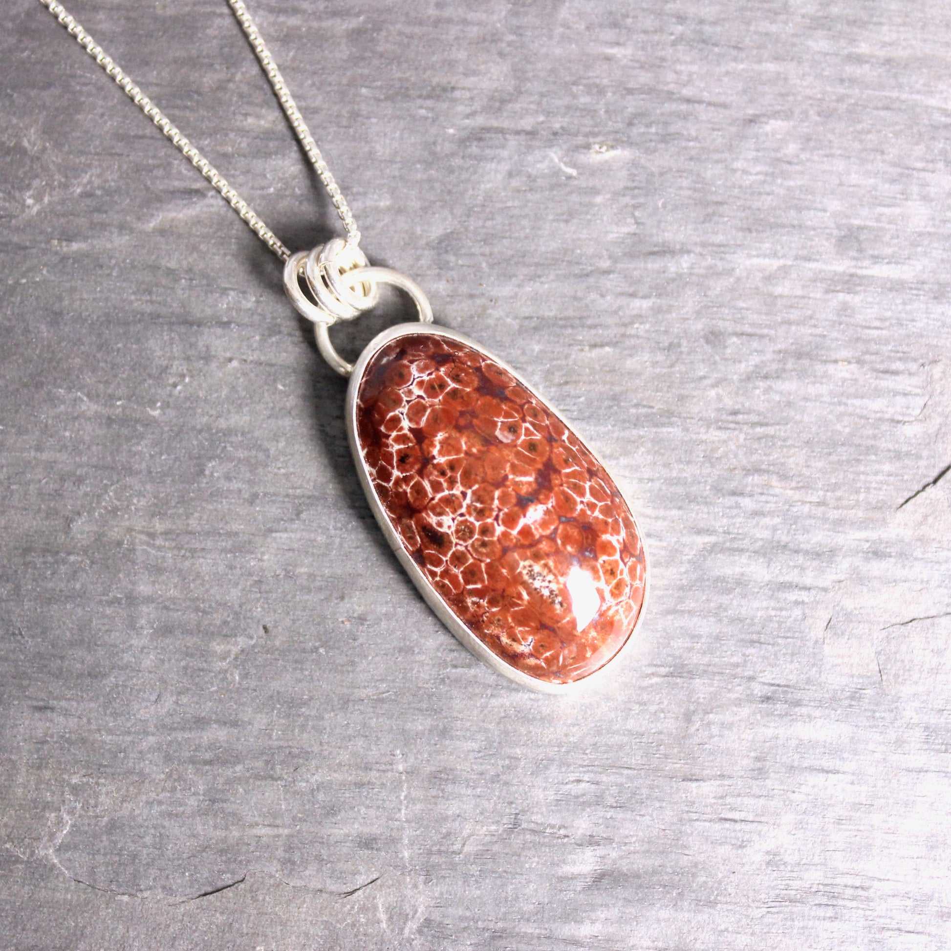 May include: A silver chain necklace with a red and white oval pendant. The pendant is made of a natural stone with a unique pattern.