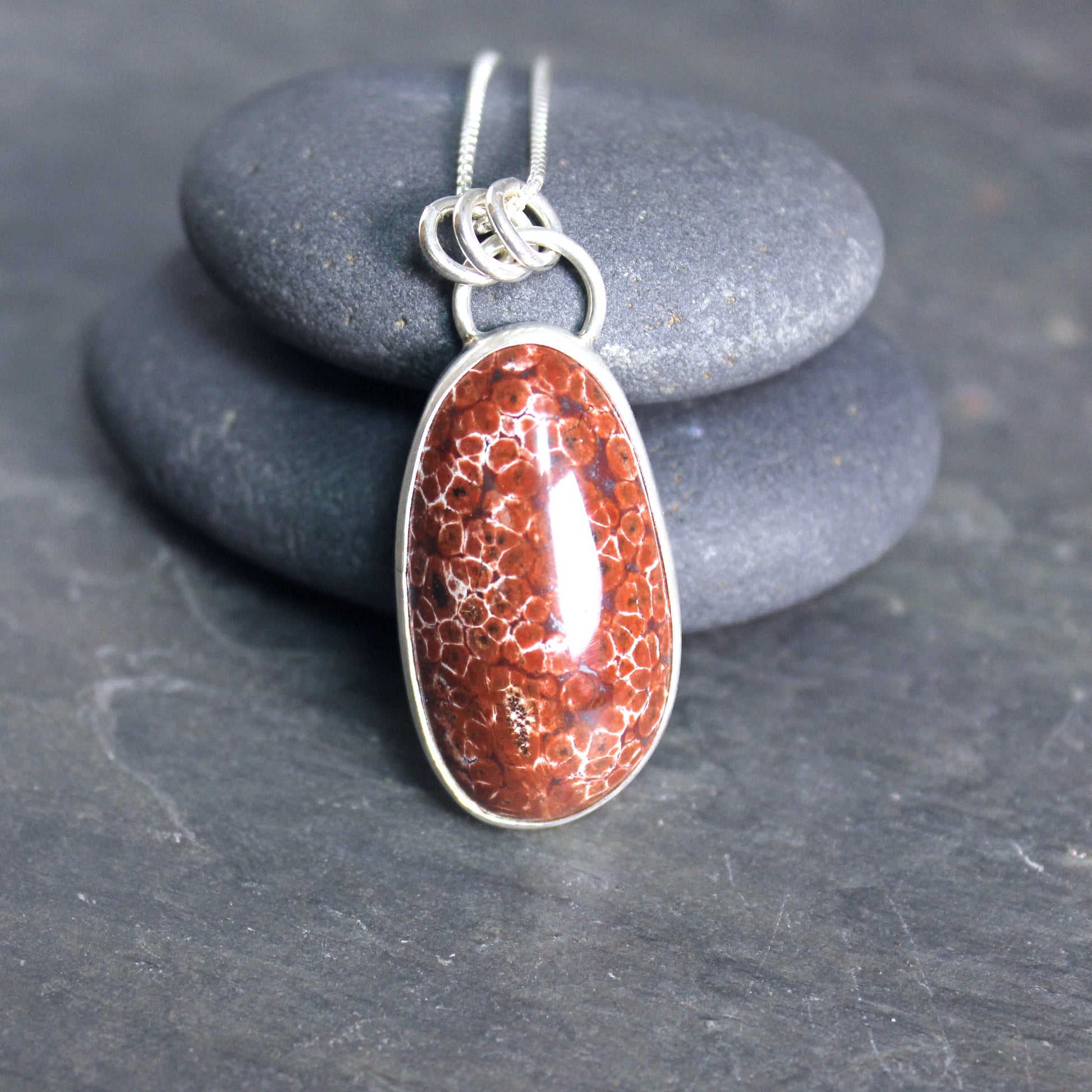 May include: A silver chain necklace with a red and white oval pendant. The pendant is made of a natural stone with a unique pattern.