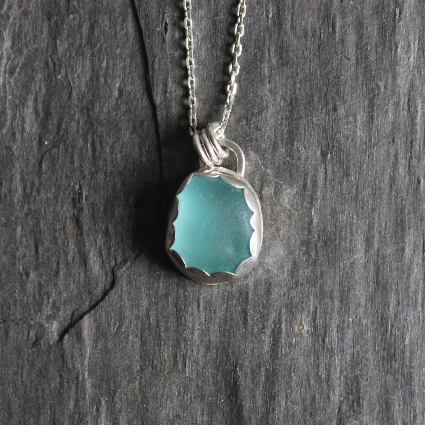 A small piece of aqua blue sea glass set in a fine and sterling silver scalloped bezel setting on a sterling silver chain. 