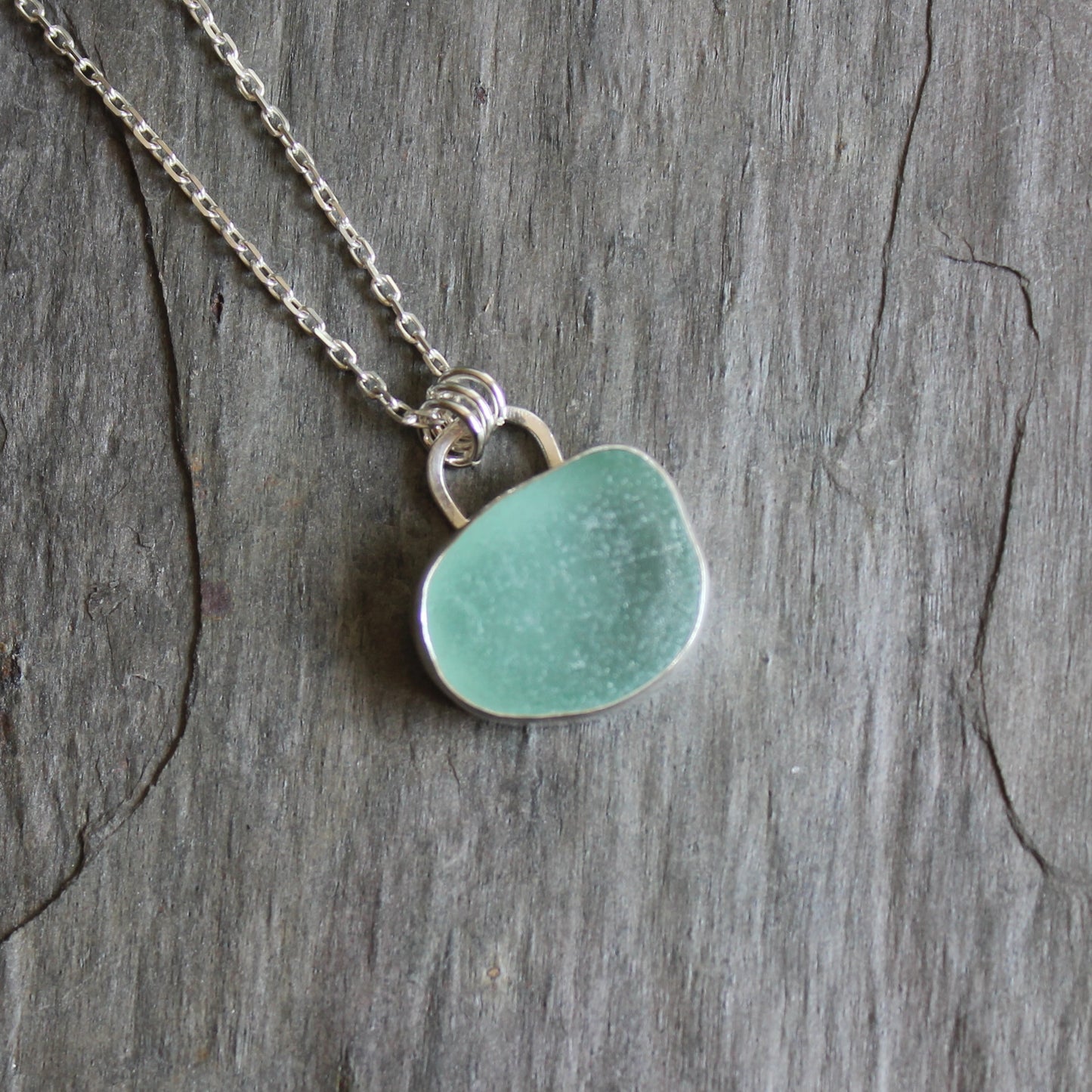 This is a large light teal piece of sea glass that has been set in a fine and sterling silver bezel setting with the back side cut out to allow light to shine through. 