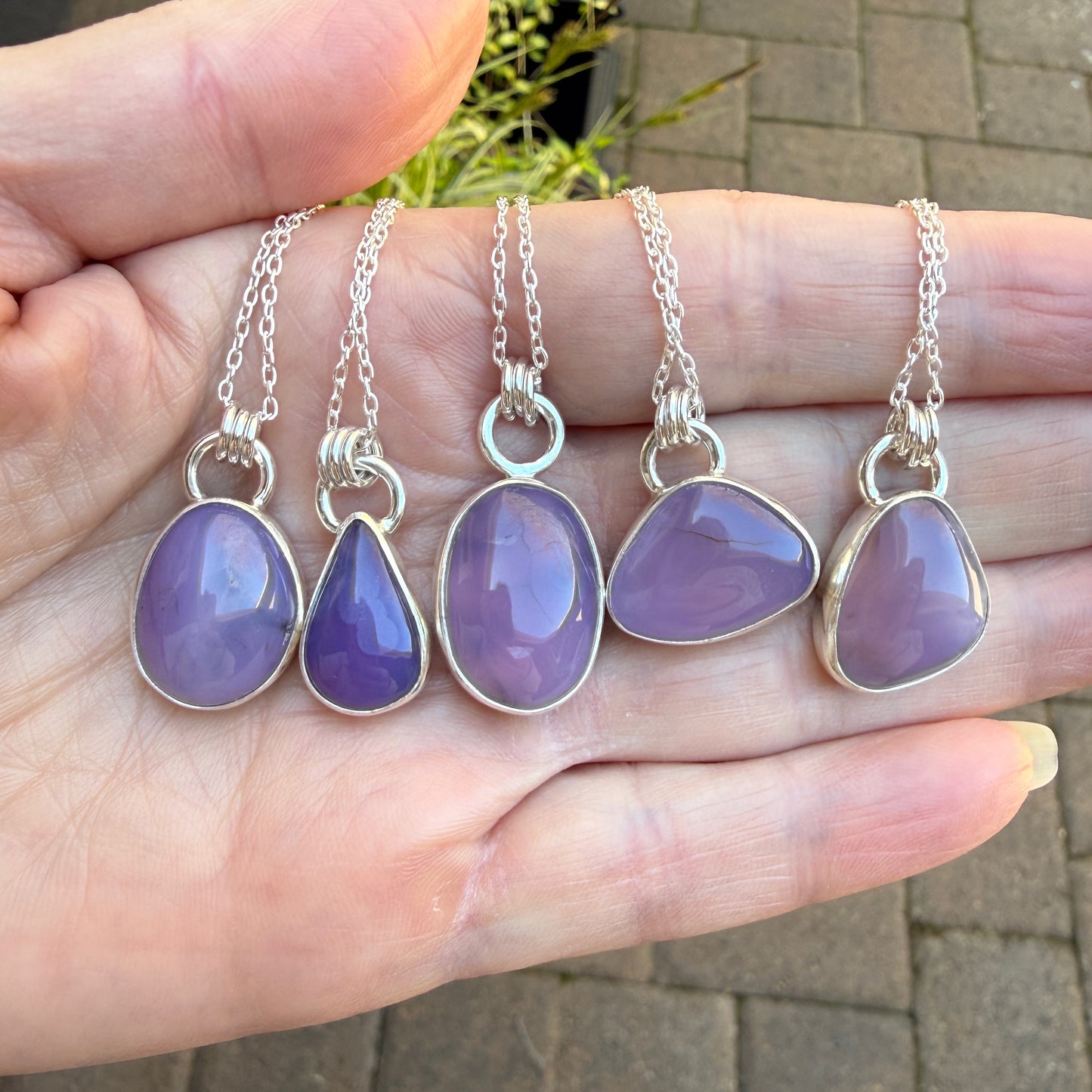Purple holley blue agate pendants on silver chains held in a hand with a blurred outdoor background.