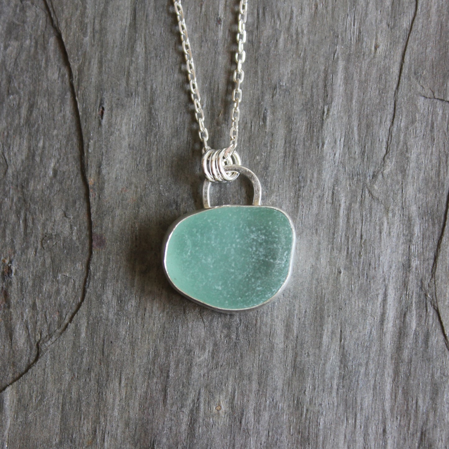 This is a large light teal piece of sea glass that has been set in a fine and sterling silver bezel setting with the back side cut out to allow light to shine through. 