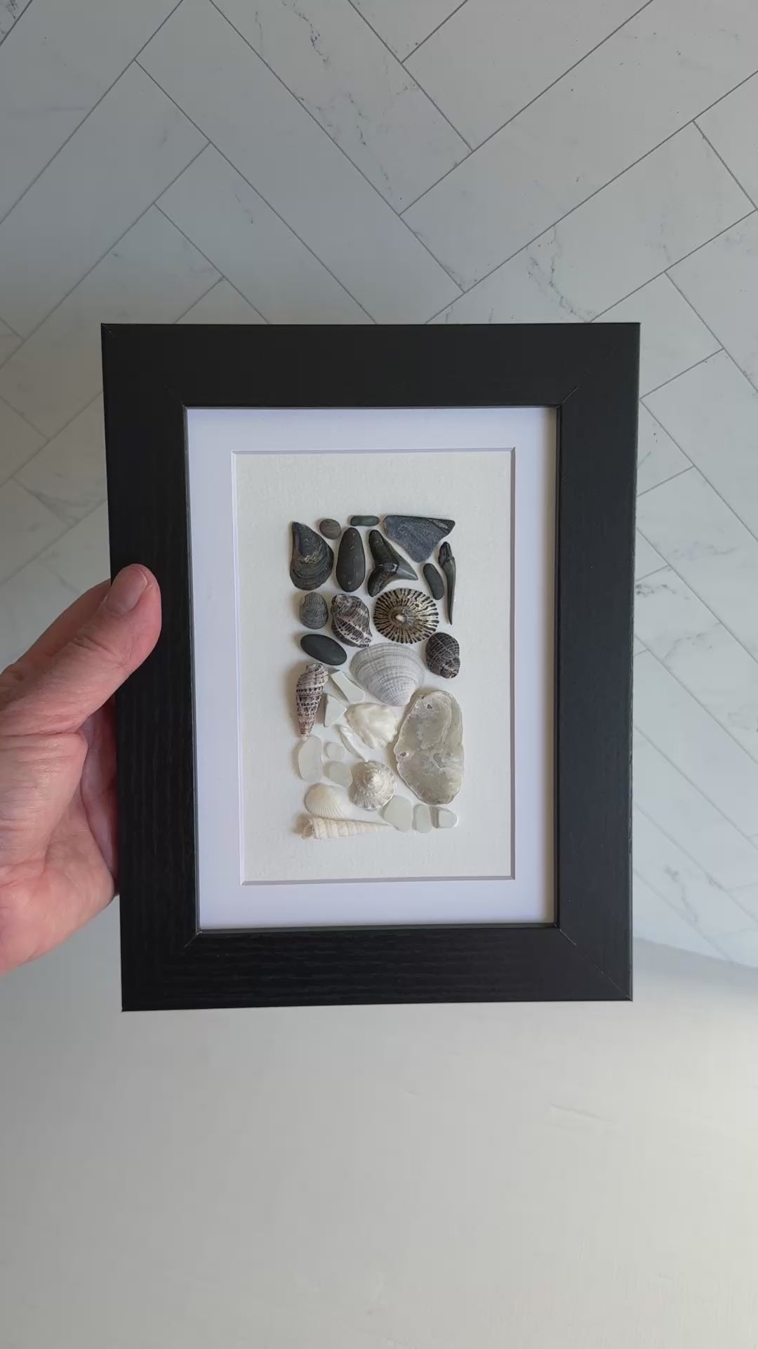Framed artwork of pebbles, shells and sea glass on a white background