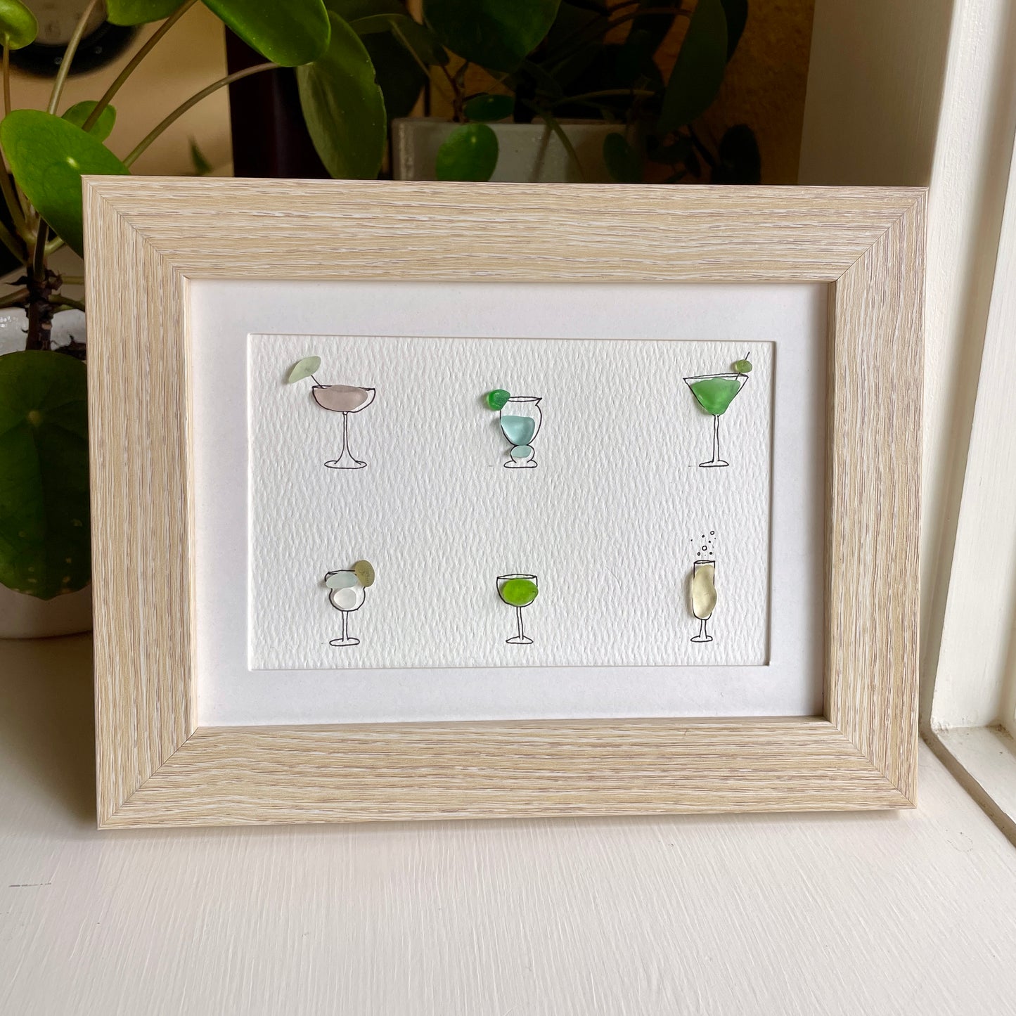 This is a framed 5 x 7 inch sea glass art picture featuring several hand-drawn cocktail and wine glasses that are filled with colorful sea glass. This item is perfect for gift giving and is ready to hang or place on a shelf.