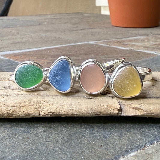 Pastel sea glass rings set in sterling silver arranged on a piece of driftwood. 