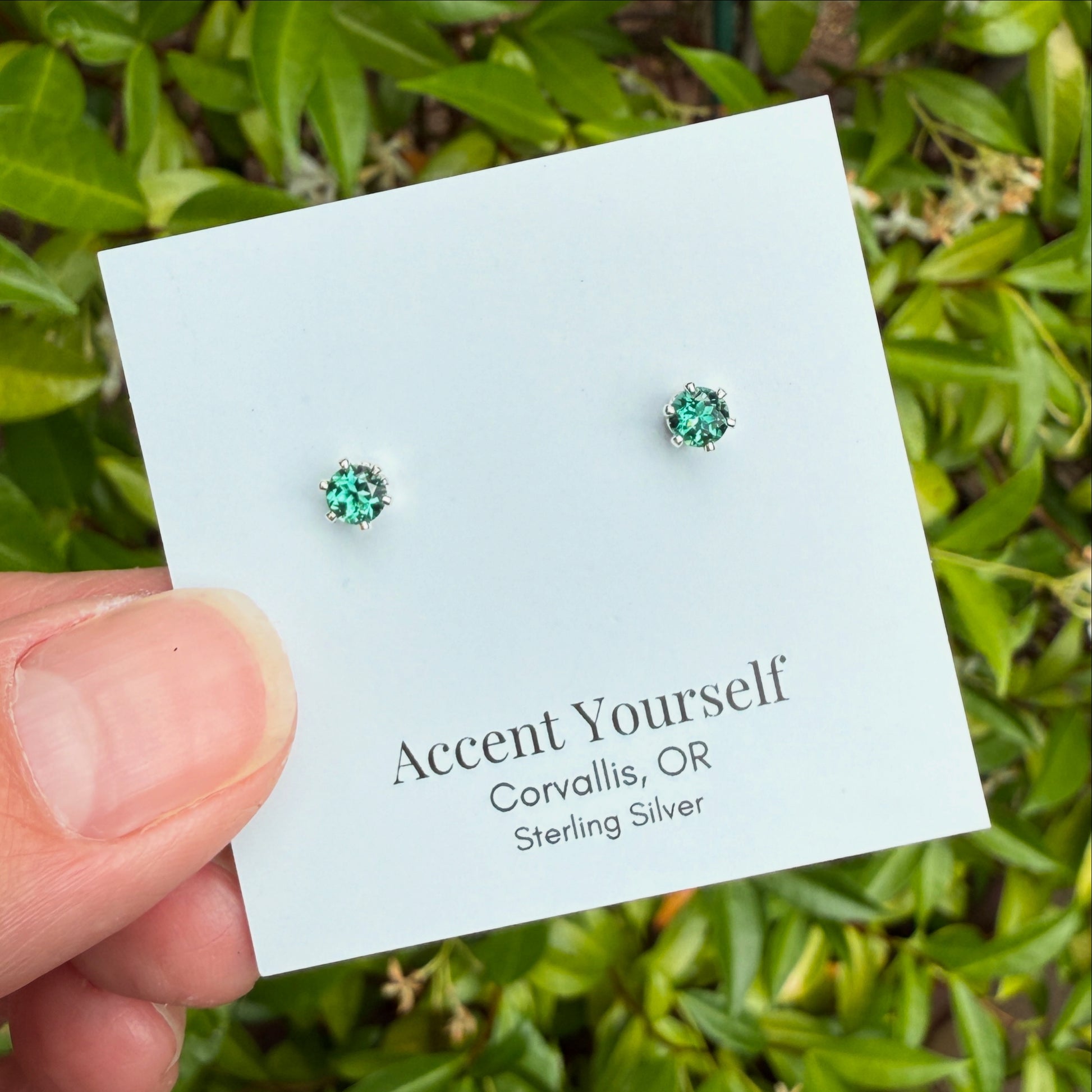 Green gemstone earrings on a 'Accent Yourself' card with a green leafy background