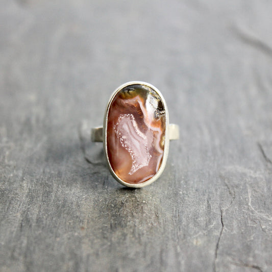 Silver ring with an oval stone on a gray surface.T his pretty ring has an oval oblong pinkish Mexican agua nueva agate cabochon set in a fine and sterling silver bezel setting on a sturdy hammered band. Size 6  Handmade by Will Macy in Corvallis, OR.