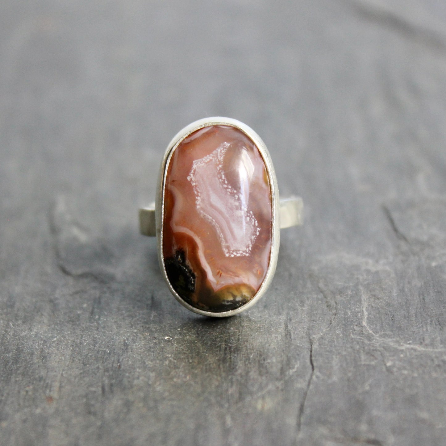 Silver ring with a large brown stone on a gray surface.This pretty ring has an oval oblong pinkish Mexican aqua nueva agate cabochon set in a fine and sterling silver bezel setting on a sturdy hammered band. Size 6  Handmade by Will Macy in Corvallis, OR.