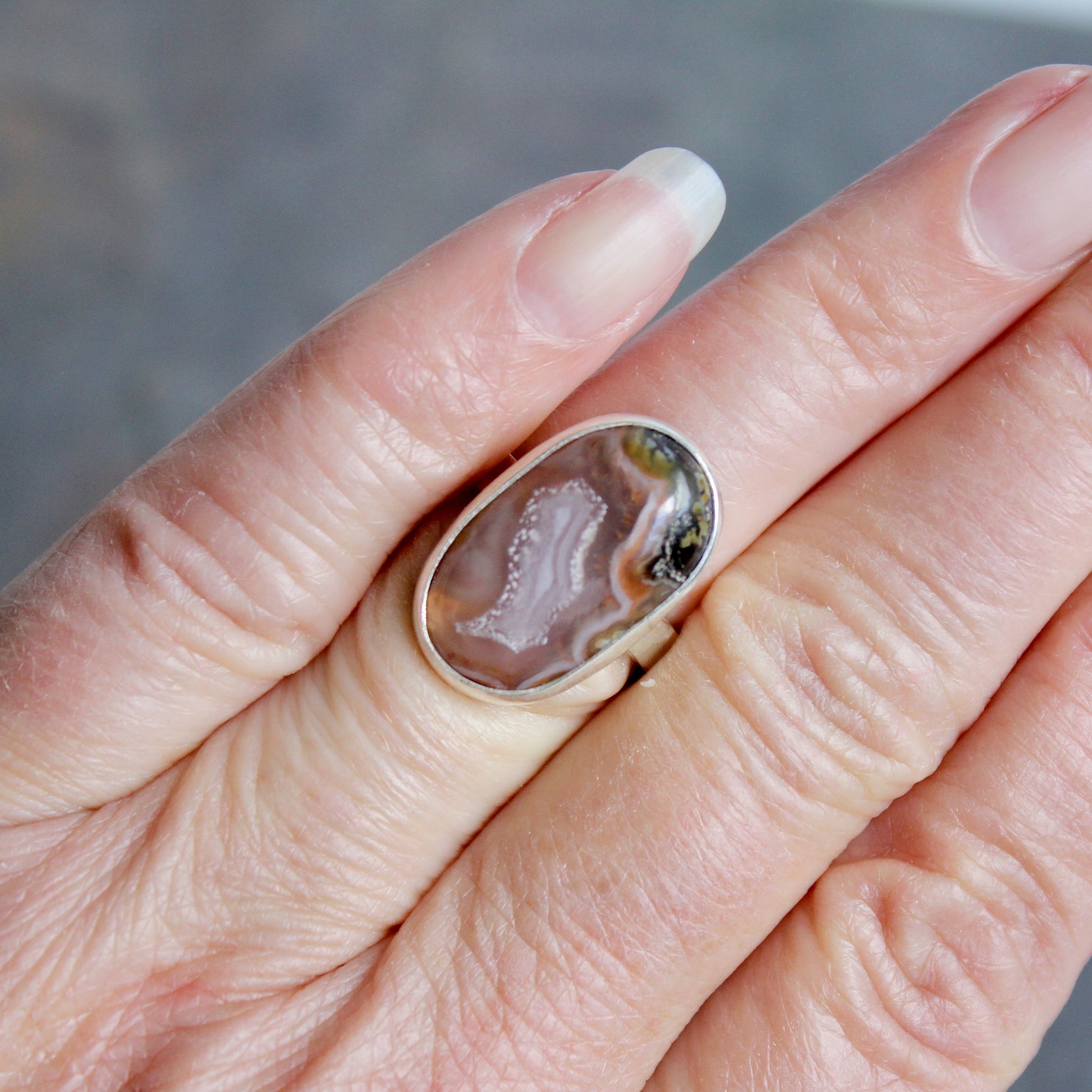 Hand wearing a ring with a large, textured stone on a neutral background. This pretty ring has an oval oblong pinkish Mexican aqua nueva agate cabochon set in a fine and sterling silver bezel setting on a sturdy hammered band. Size 6  Handmade by Will Macy in Corvallis, OR.
