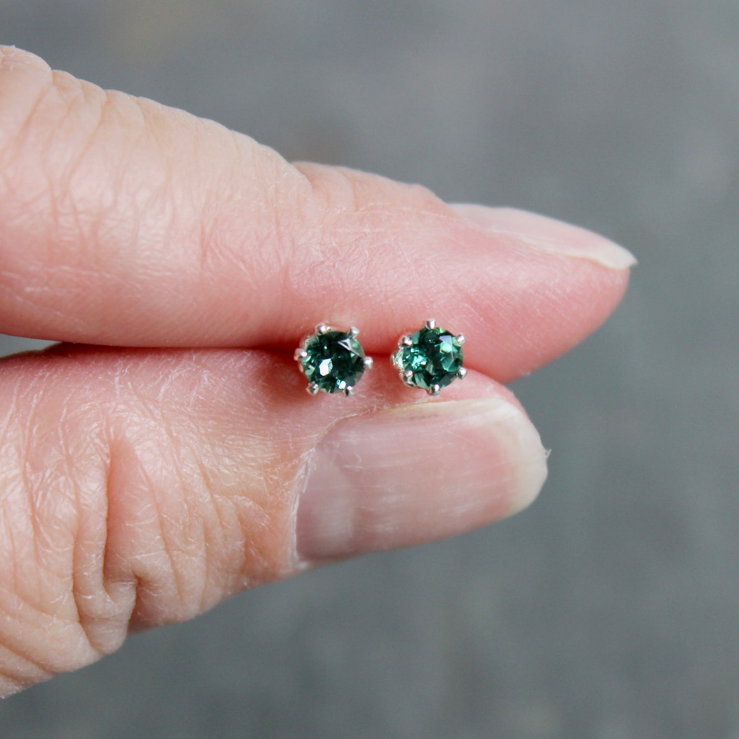 Green gemstone stud earrings held between fingers against a blurred background
