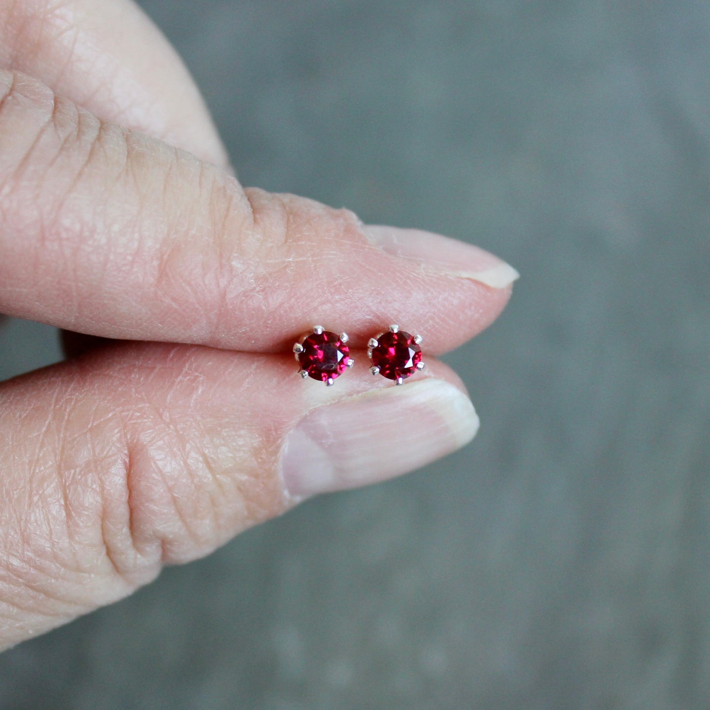 Red gemstone stud earrings held between fingers against a neutral background