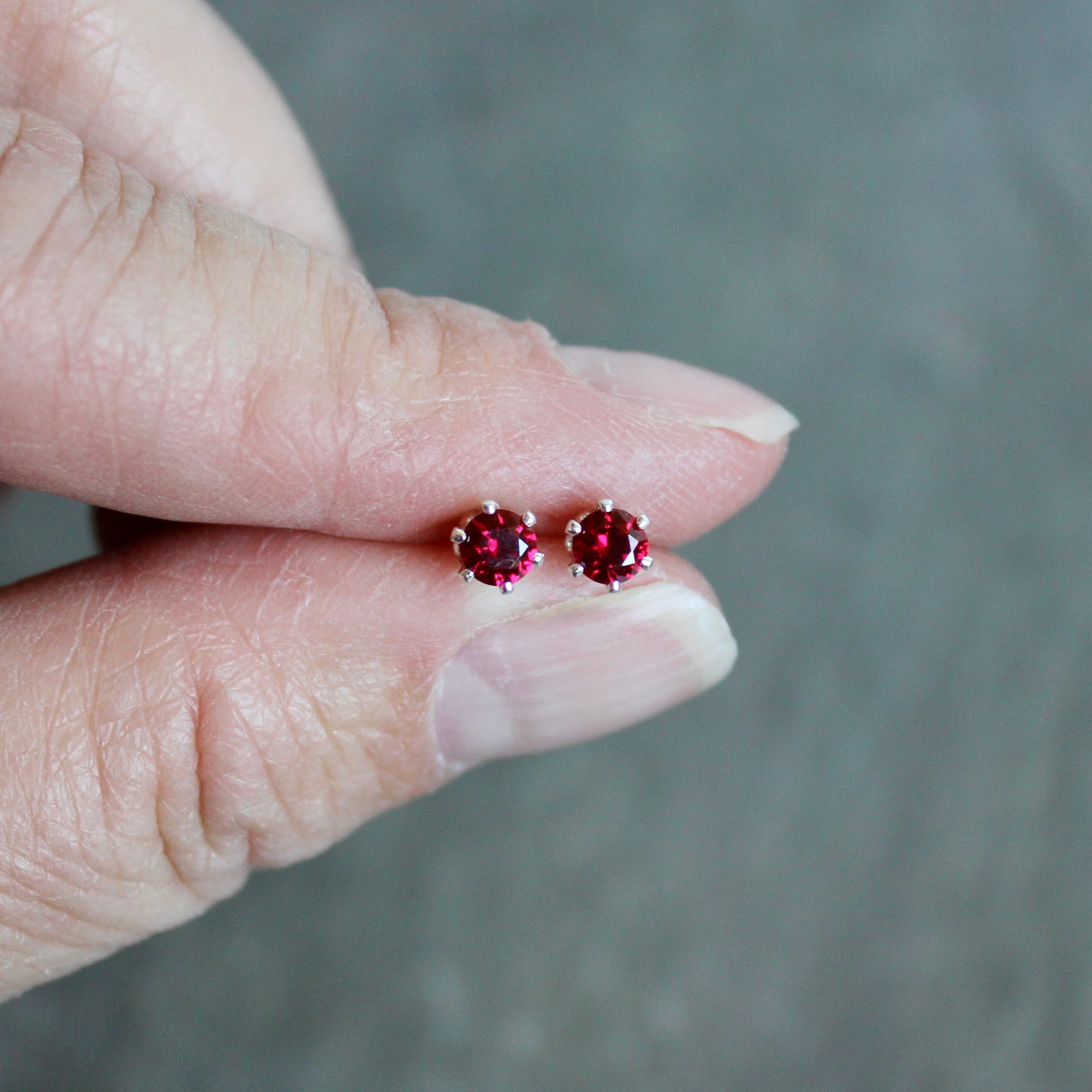Red gemstone stud earrings held between fingers against a neutral background