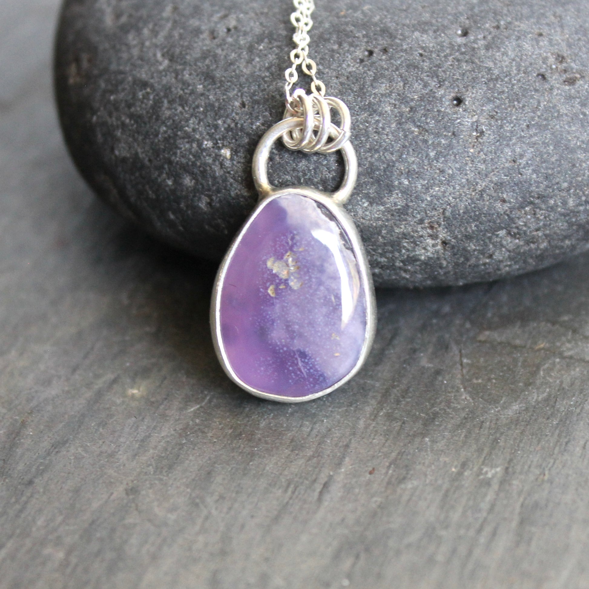 Purple teardrop pendant on a silver chain against a gray stone background. A holley blue agate cabochon set in a fine and sterling silver bezel setting on a sturdy silver chain. 