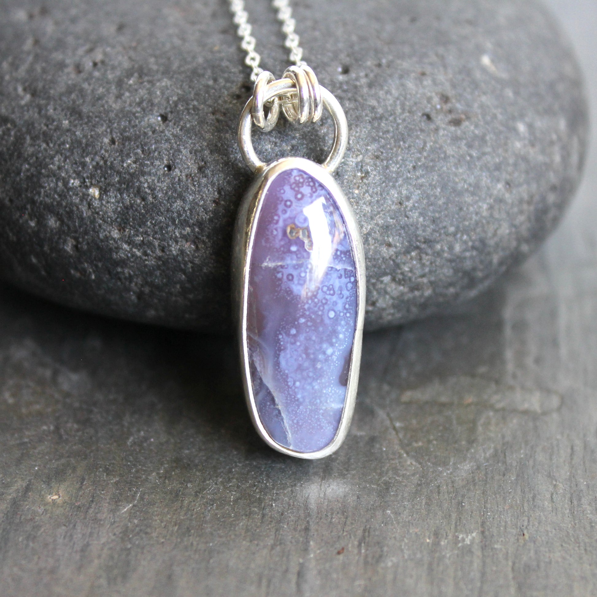 Purple pendant necklace on a gray stone surface. A long holley blue agate cabochon set in a fine and sterling silver bezel setting on a sturdy silver chain. 