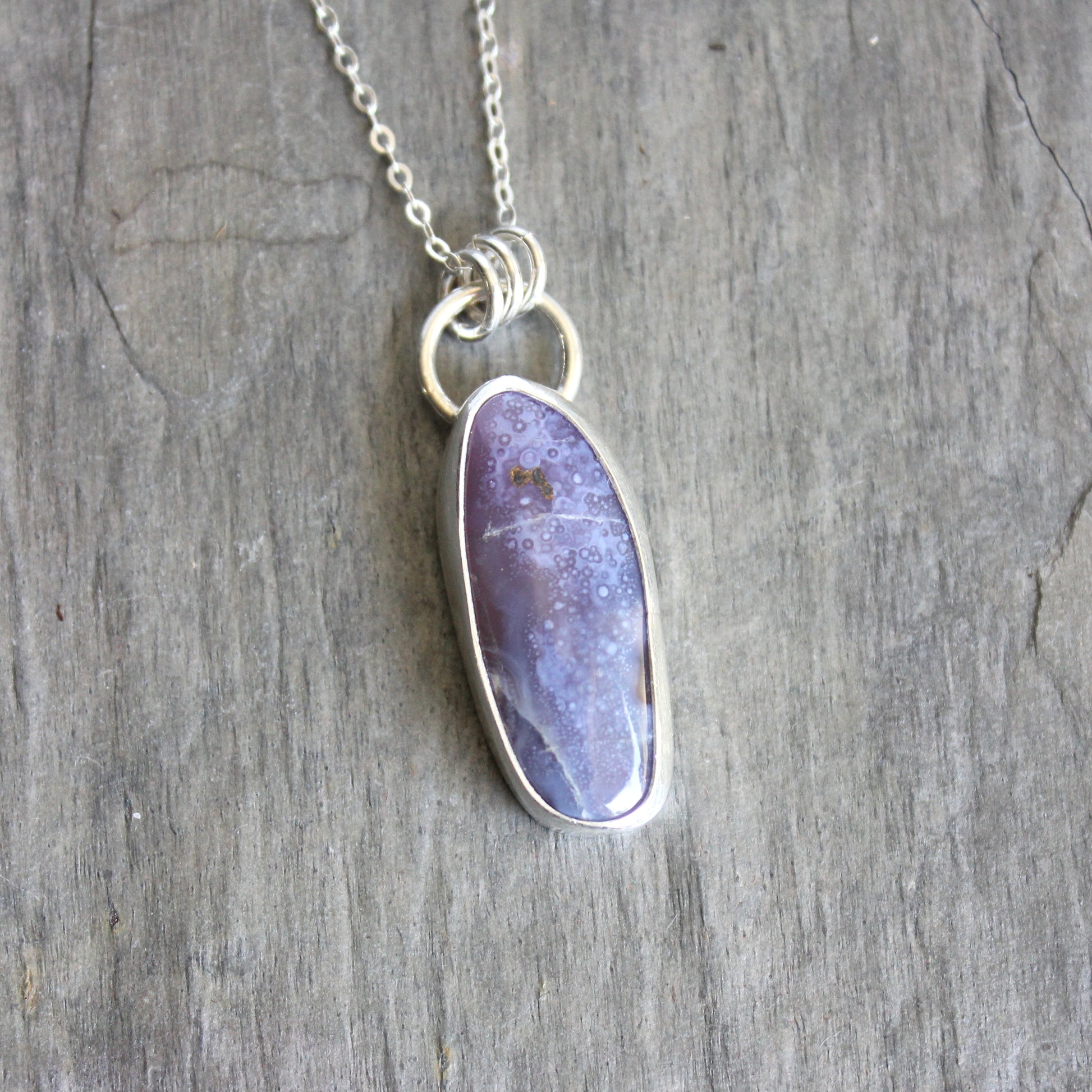 Necklace with a purple oval pendant on a wooden surface. A long holley blue agate cabochon set in a fine and sterling silver bezel setting on a sturdy silver chain. 