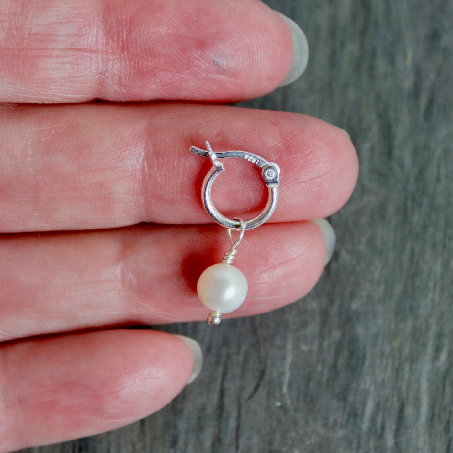 Silver hoop earring with a pearl on a hand against a gray background. These locking hoops are approximately 10mm in diameter with a removeable freshwater pearl drop.