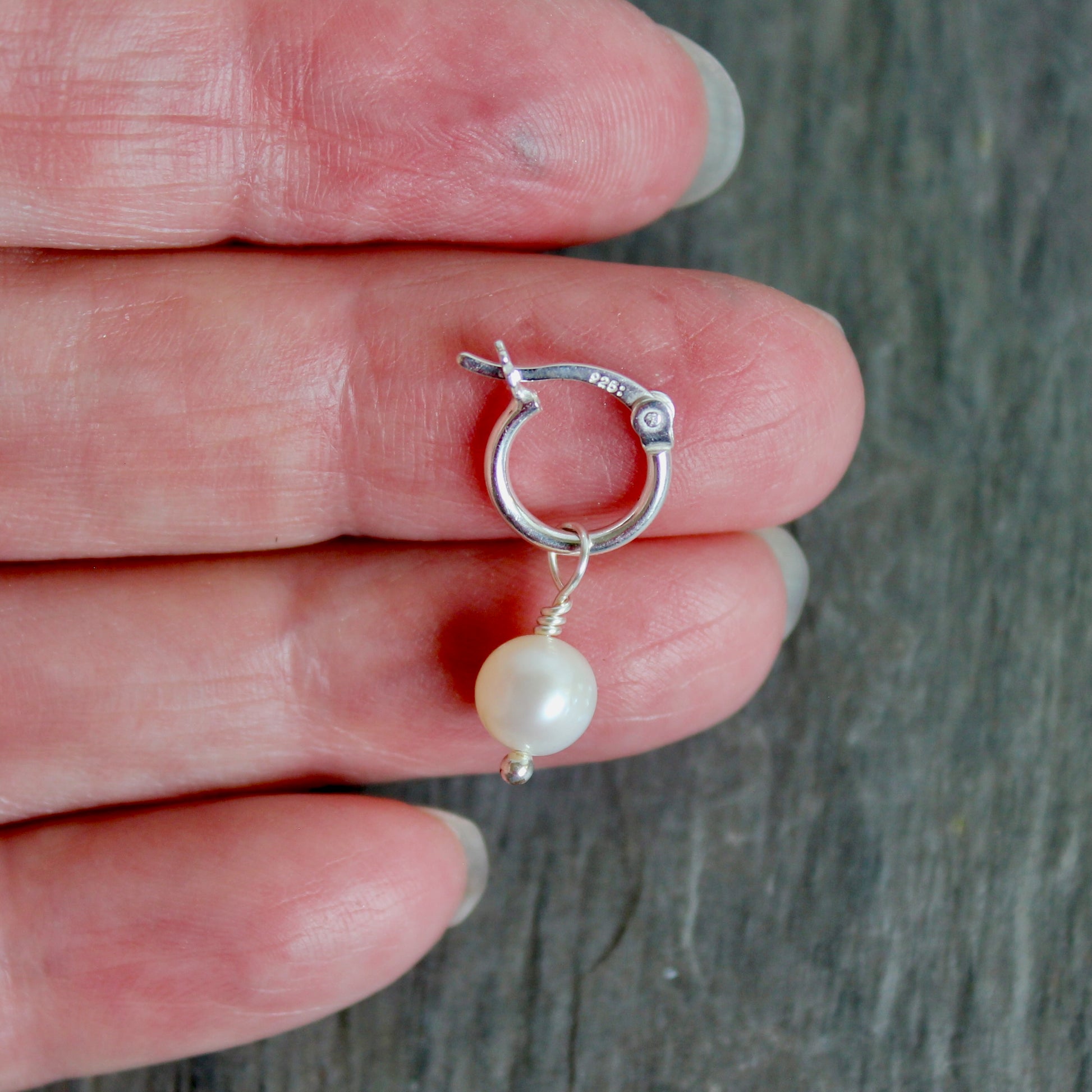 Silver hoop earring with a pearl on a hand against a gray background. These locking hoops are approximately 10mm in diameter with a removeable freshwater pearl drop.