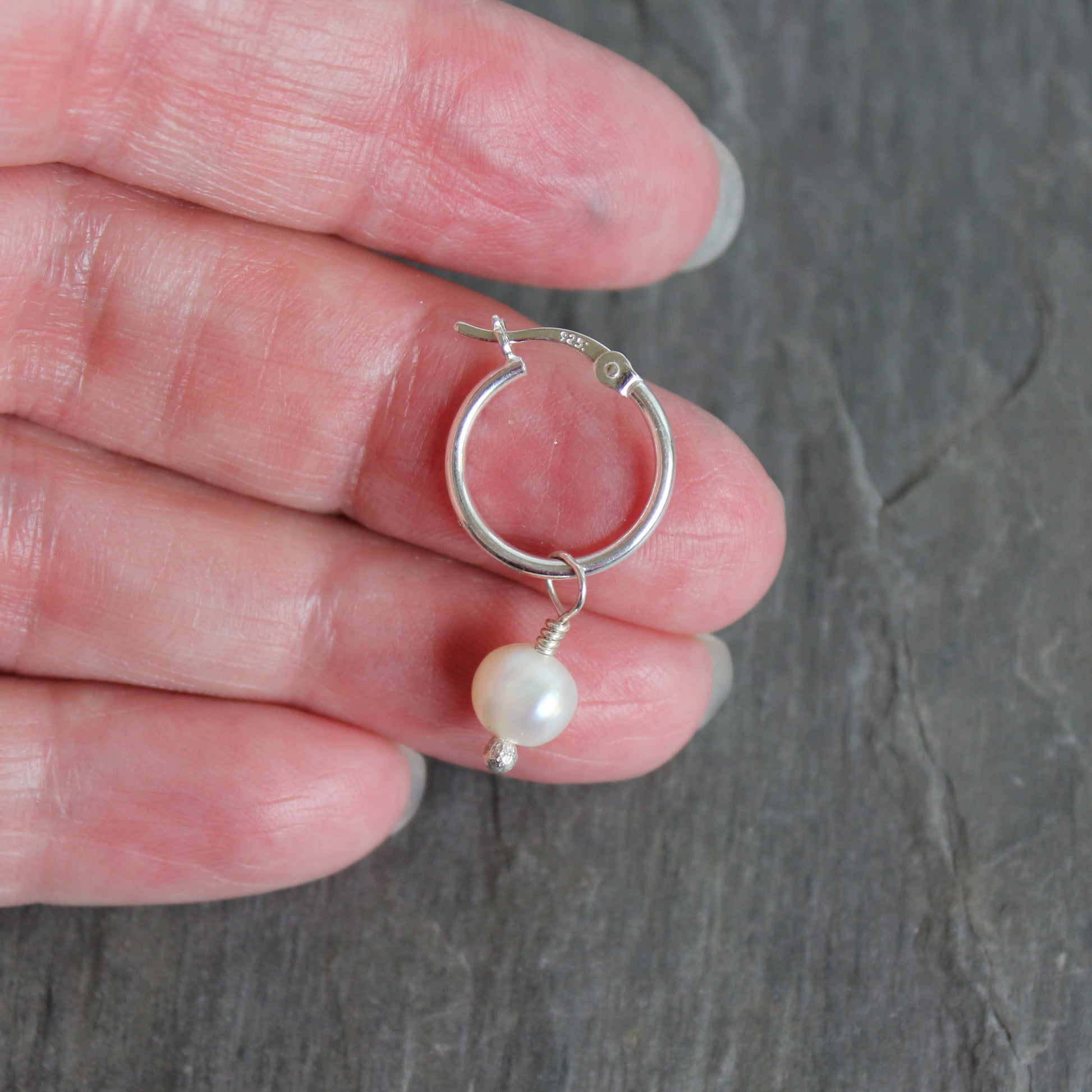Silver hoop earring with a pearl on a hand against a gray background. These locking hoops are approximately 15mm in diameter with a removeable freshwater pearl drop.