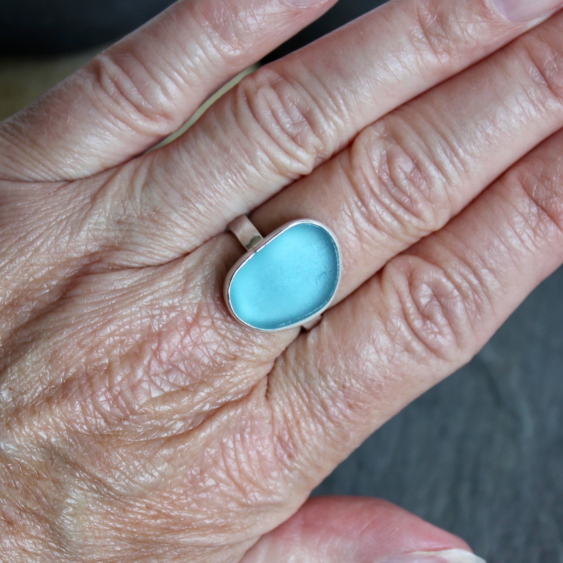 Close-up of a hand wearing a blue ring with a blurred background.This ring has a large piece of aqua turquoise blue sea glass set in a fine and sterling silver bezel on a sturdy hammered band. Size 8 1/2 Handmade by Barb Macy in Corvallis, Oregon.