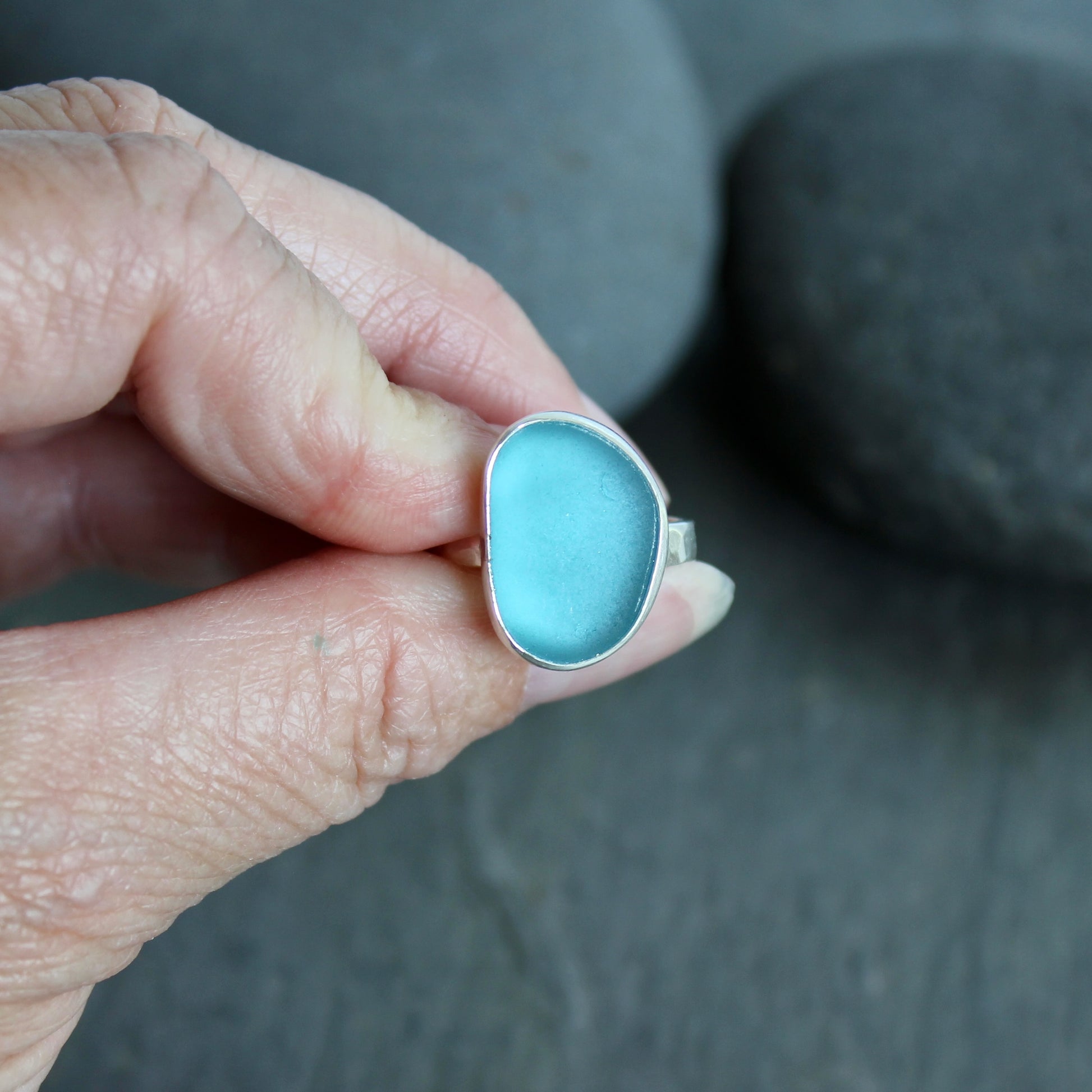 Hand holding a turquoise stone with silver setting against a dark background.This ring has a large piece of aqua turquoise blue sea glass set in a fine and sterling silver bezel on a sturdy hammered band. Size 8 1/2 Handmade by Barb Macy in Corvallis, Oregon.