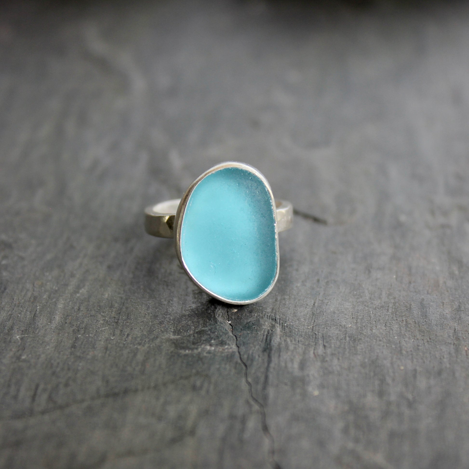 Blue ring with silver band on a dark stone surface. This ring has a large piece of aqua turquoise blue sea glass set in a fine and sterling silver bezel on a sturdy hammered band. Size 8 1/2 Handmade by Barb Macy in Corvallis, Oregon.