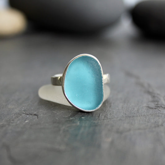 Silver ring with a blue stone on a dark wooden surface.This ring has a large piece of aqua turquoise blue sea glass set in a fine and sterling silver bezel on a sturdy hammered band. Size 8 1/2 Handmade by Barb Macy in Corvallis, Oregon.