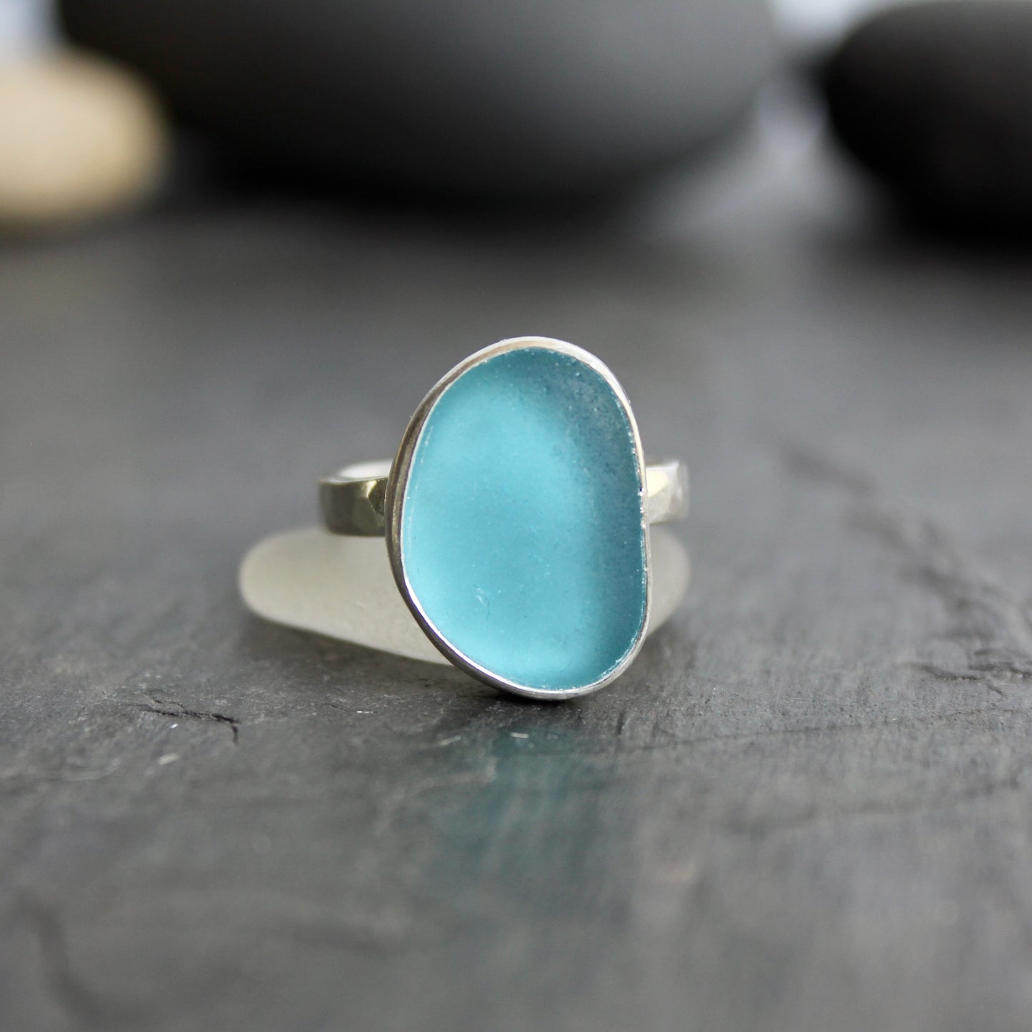 Silver ring with a blue stone on a dark wooden surface.This ring has a large piece of aqua turquoise blue sea glass set in a fine and sterling silver bezel on a sturdy hammered band. Size 8 1/2 Handmade by Barb Macy in Corvallis, Oregon.